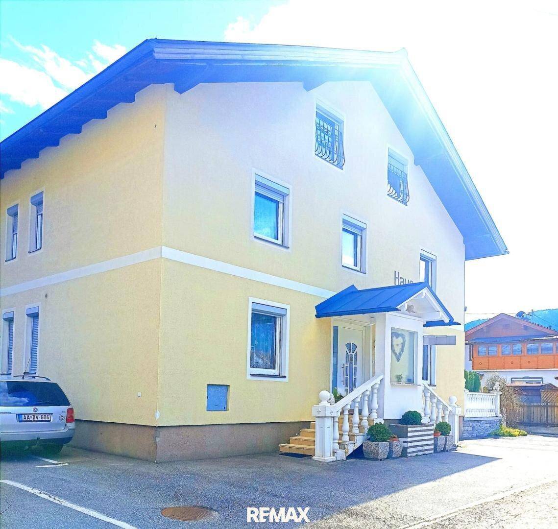 Gelbes Haus mit blauem Dach und Eingangsbereich, umgeben von viel Grün und Sonnenschein.