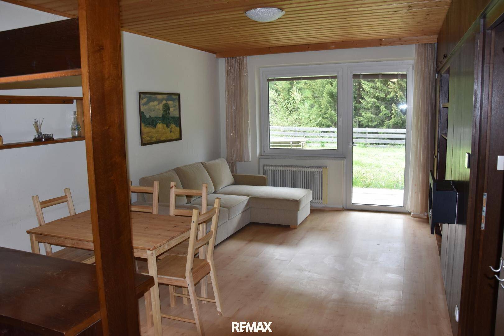 Heller Wohnbereich mit Sofa, Holzboden und großem Fenster mit Blick ins Grüne.