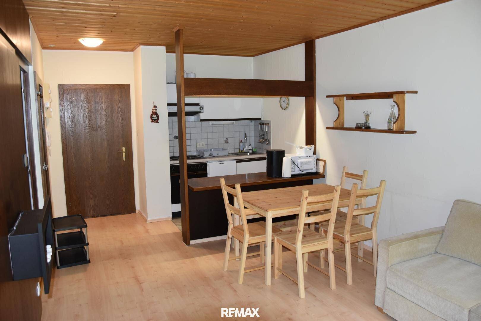 Gemütlicher Wohn-Essbereich mit offener Küchenzeile, Holztisch und Sofa unter einer Holzdecke.