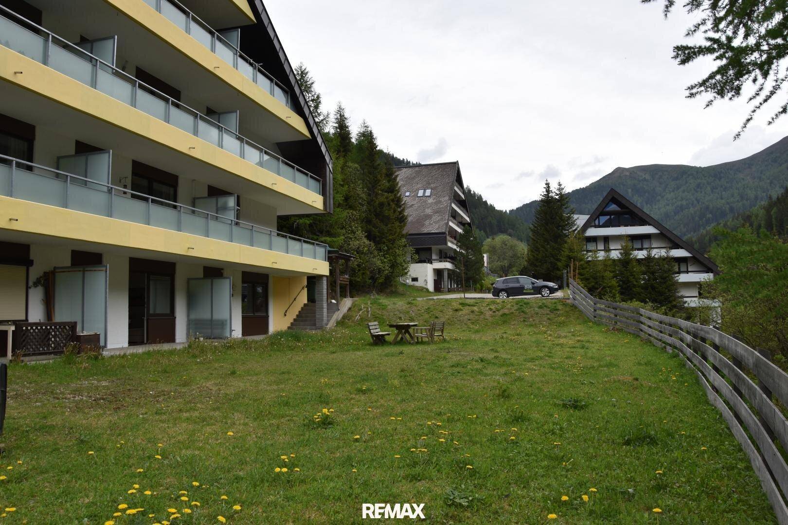 Gebäudekomplex in den Bergen mit Balkonen, umgeben von Wiesen und Nadelbäumen unter bewölktem Himmel.