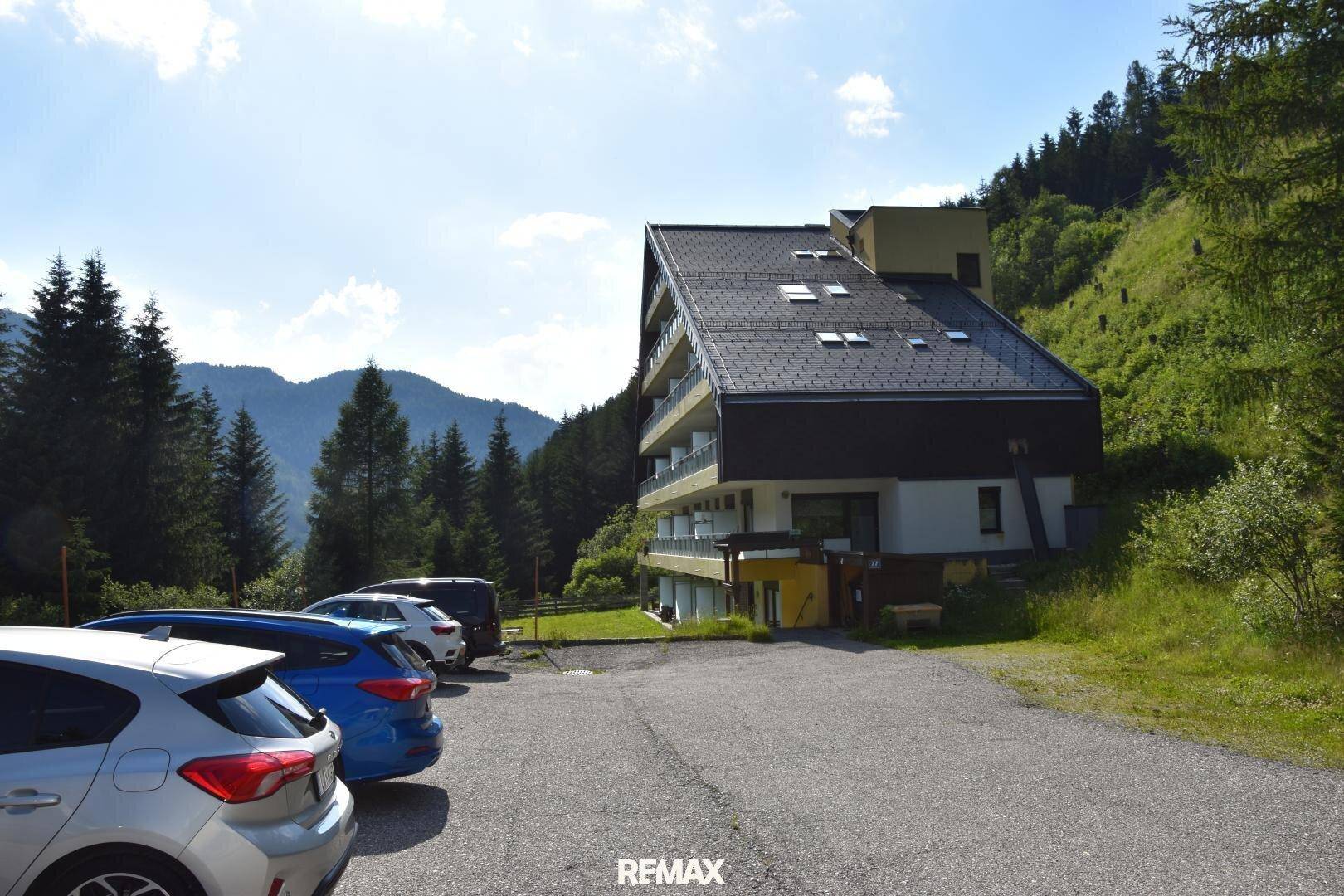 Modernes Gebäude mit schrägem Dach und Balkonen, umgeben von Bergen und Wald, mit Parkplätzen davor.