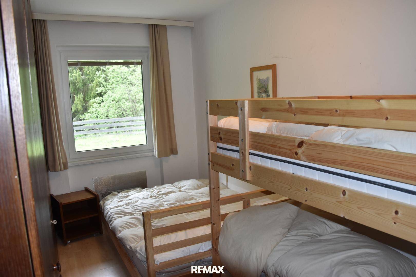Schlafzimmer mit Etagenbett und Einzelbett, Holzelementen und Fenster mit Blick ins Grüne.