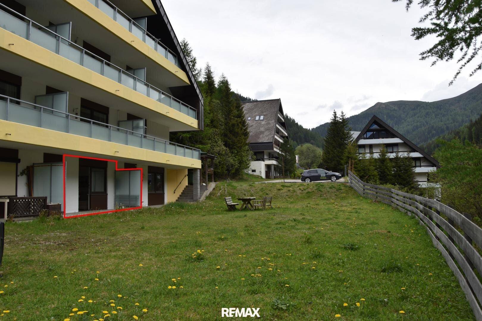 Gebäudekomplex in alpiner Umgebung mit markiertem Erdgeschossbereich, umgeben von Wiesen und Bergen.