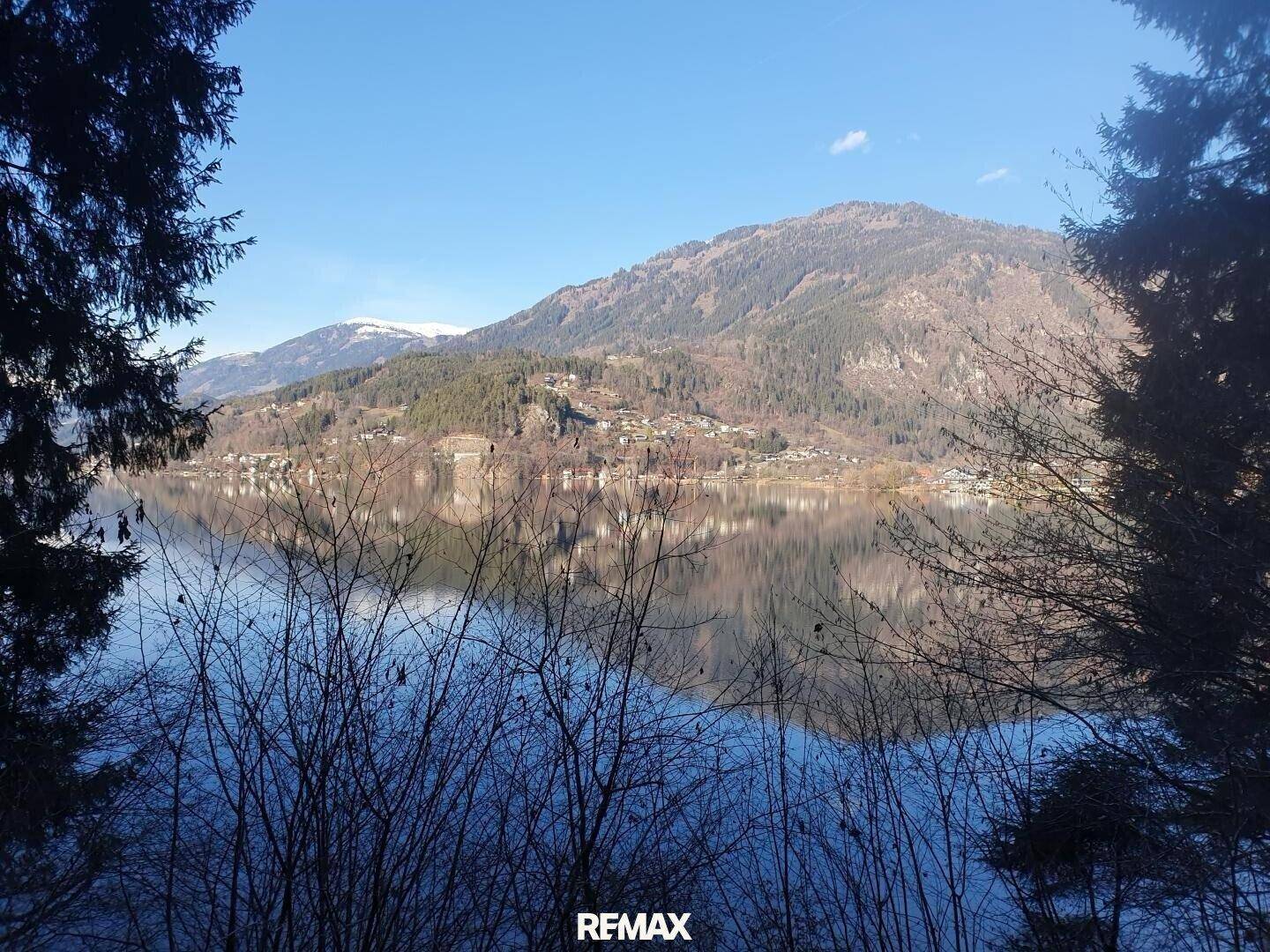 Blick auf einen See durch kahle Äste, mit einer Siedlung und Bergen am gegenüberliegenden Ufer.