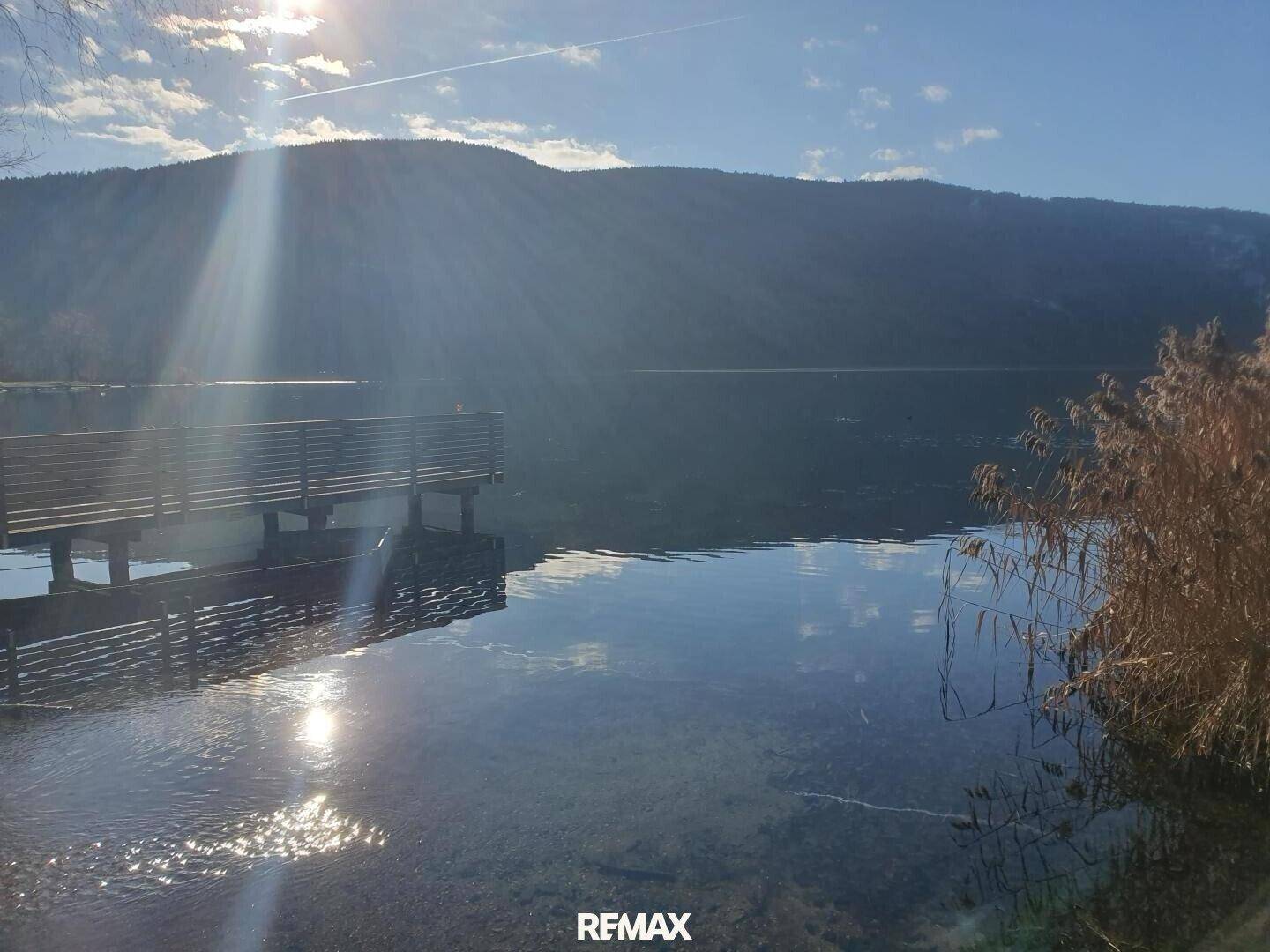 Ruhiger See mit einem Holzsteg und Schilf am Ufer, die Sonne spiegelt sich im Wasser.