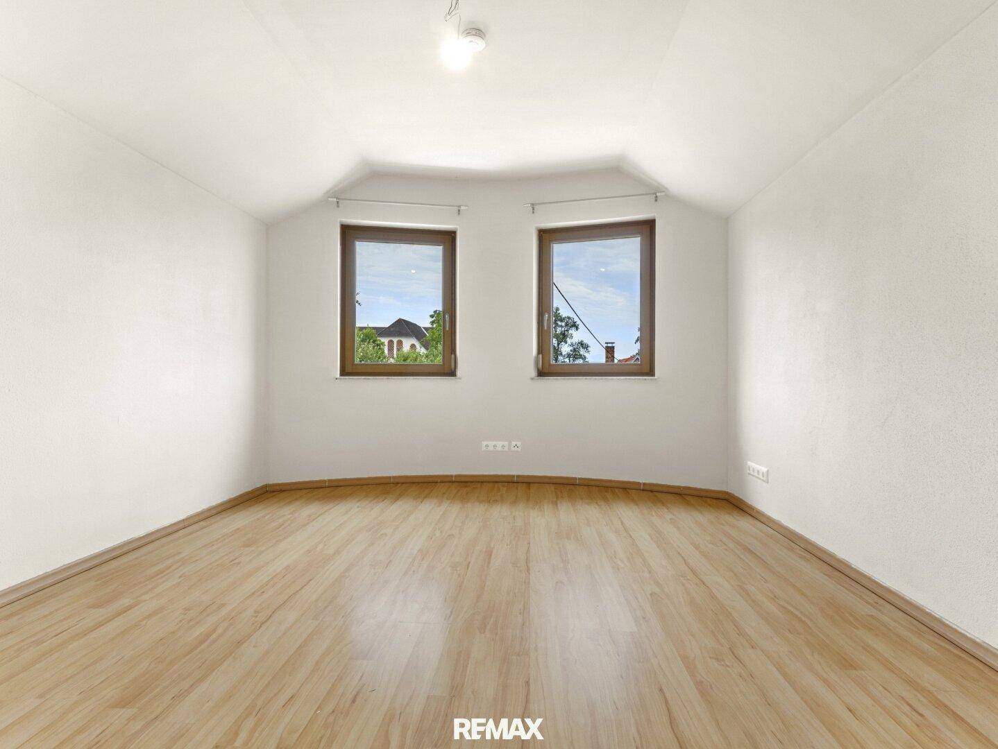 Leeres Zimmer mit Holzboden, weißen Wänden und zwei Fenstern mit Blick nach draußen.