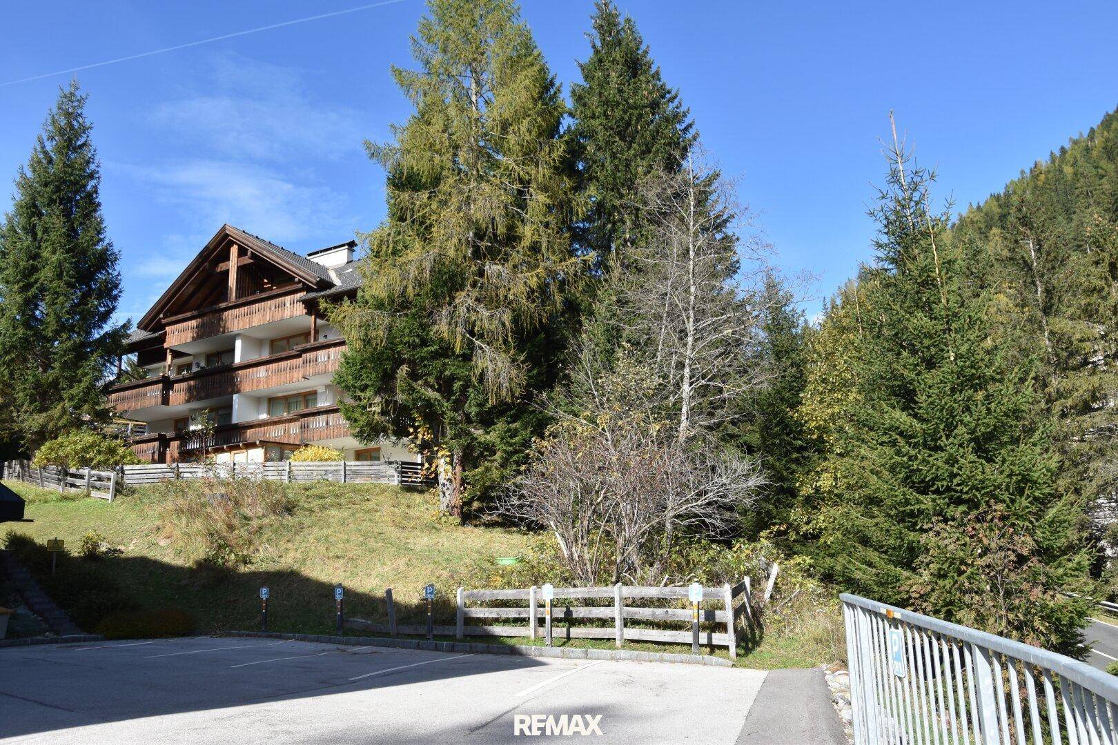 Mehrfamilienhaus im alpinen Stil mit Holzbalkonen, umgeben von hohen Bäumen und Bergen.
