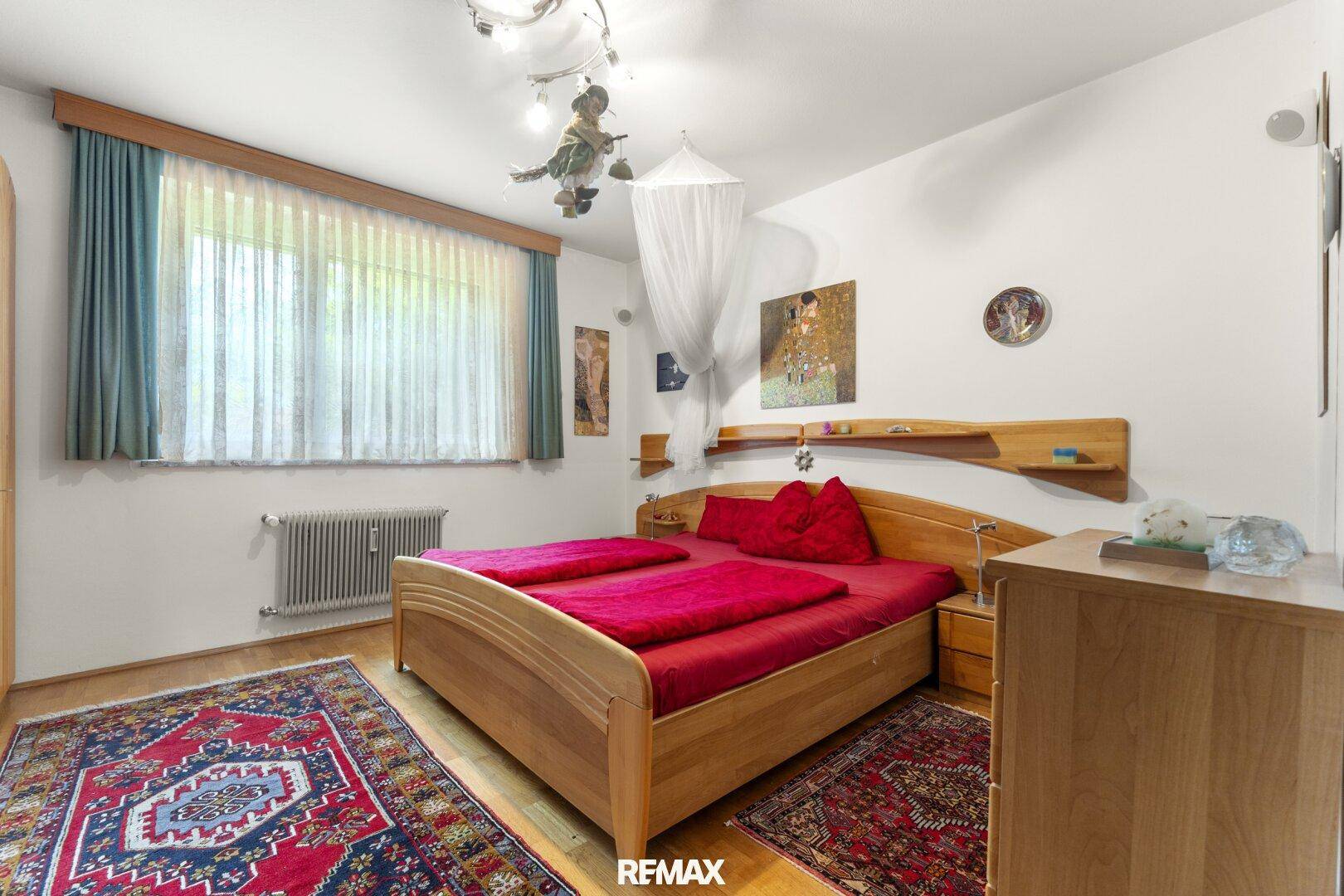 Schlafzimmer mit Doppelbett, roten Akzenten, Teppichen und großem Fenster.