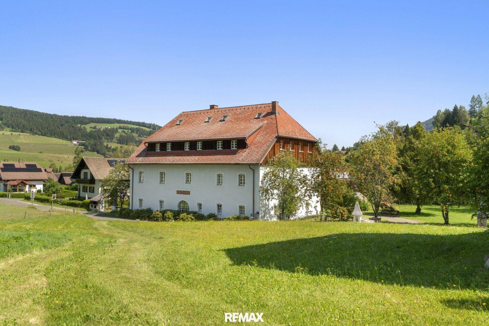 Historisches Gebäude mit weißer Fassade und rotem Ziegeldach, eingebettet in eine grüne Hügellandschaft.
