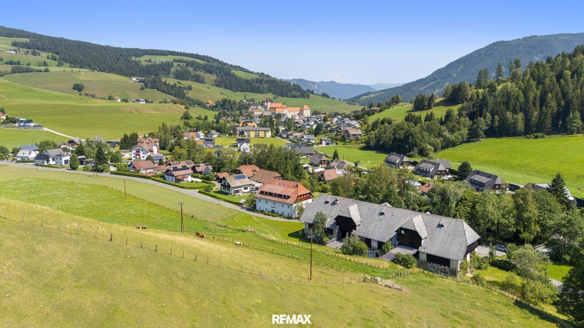 Panoramablick auf ein malerisches Dorf in einem grünen Tal, umgeben von sanften Hügeln und Wäldern.