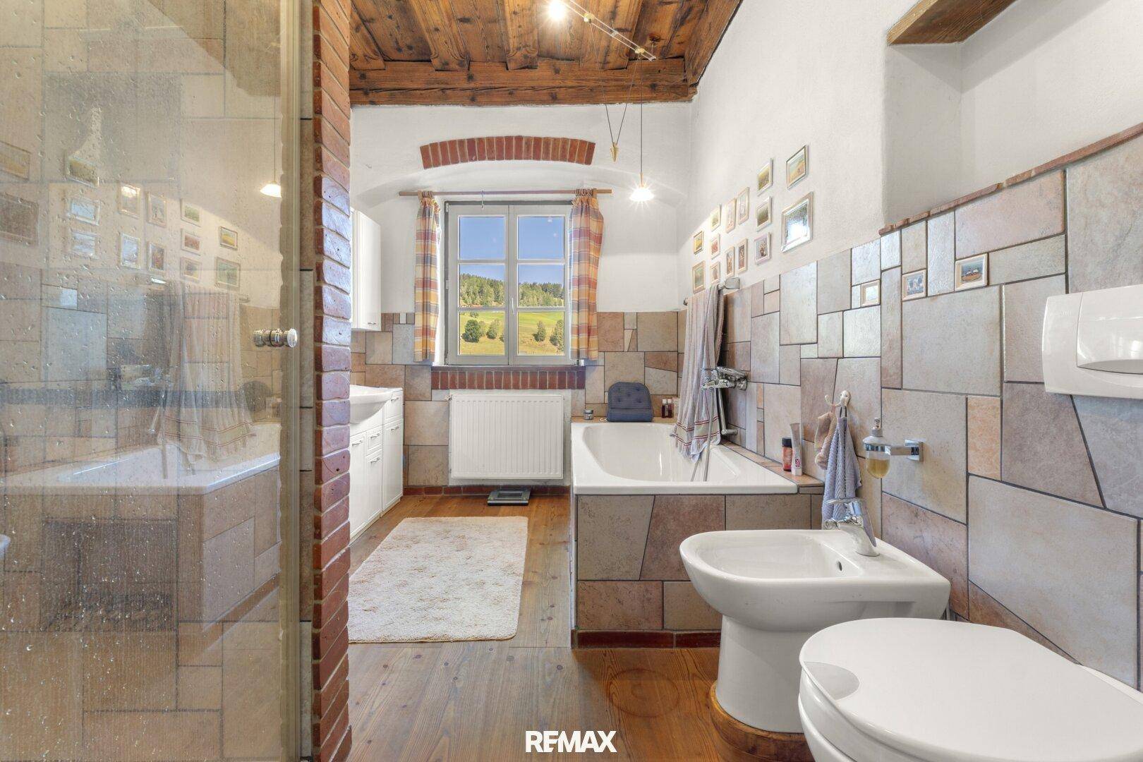 Geräumiges Badezimmer mit Badewanne, Dusche und Fliesen in Erdtönen, sowie Holzbalkendecke.