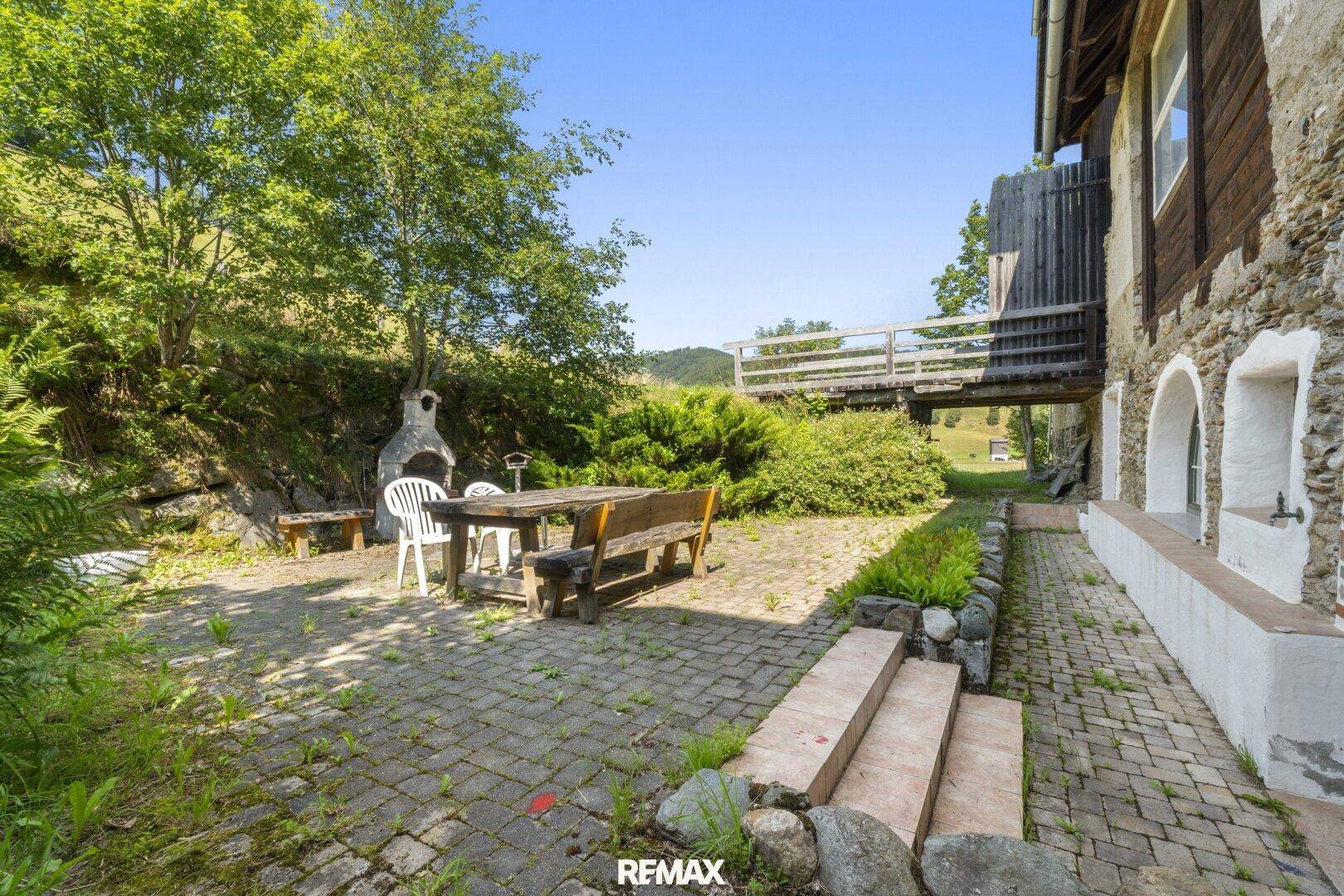 Gepflasterte Terrasse mit Sitzgelegenheiten und Grillplatz, umgeben von Bäumen und Sträuchern.