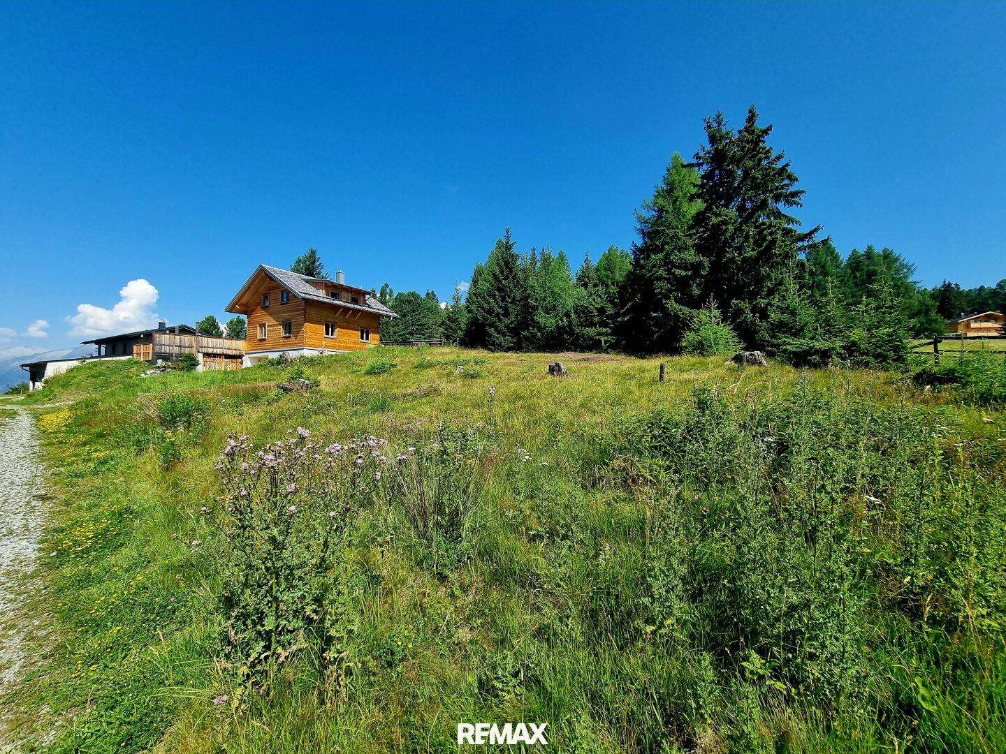 Hügelige Wiesenlandschaft mit einem charmanten Holzhaus unter strahlend blauem Himmel und viel Grün.