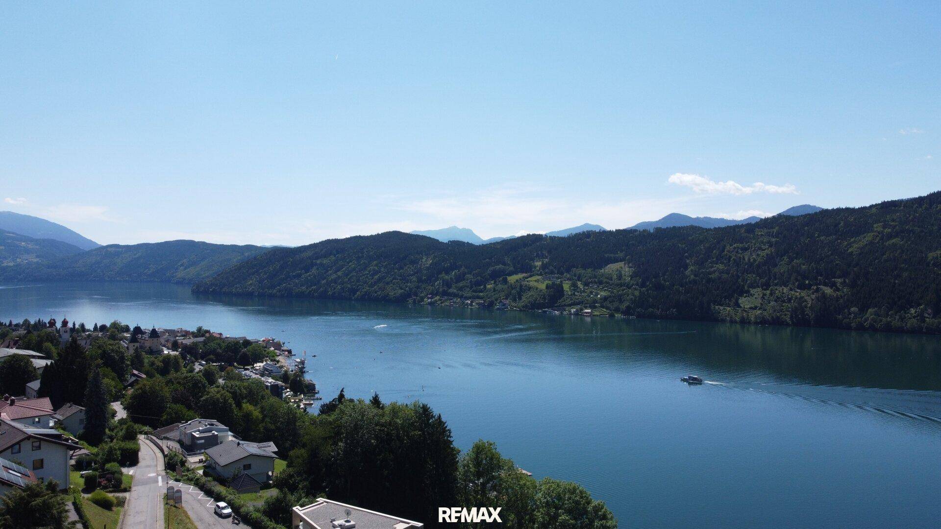 Weitläufige Luftaufnahme eines Dorfes am Seeufer mit Häusern und Bergen im Hintergrund.