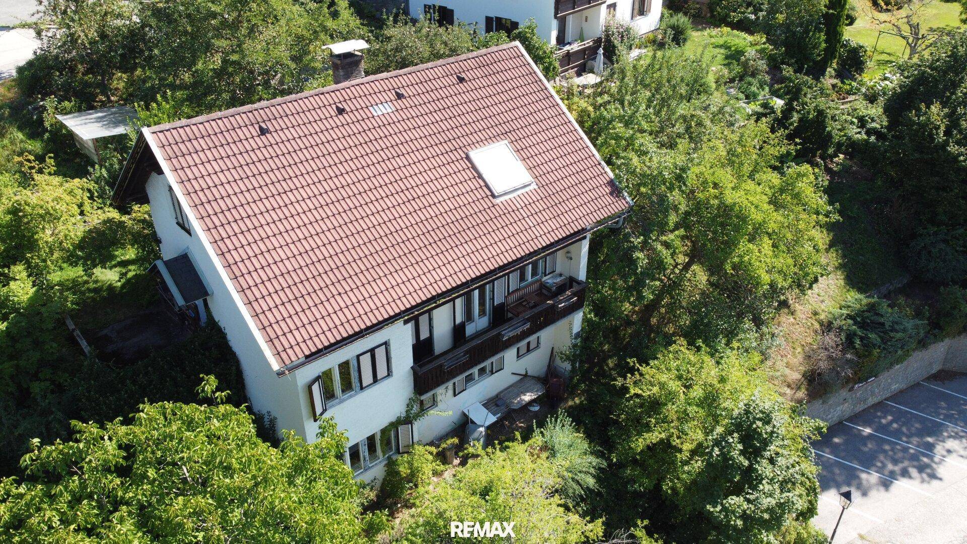 Das Haus mit rotem Ziegeldach und weißer Fassade, umgeben von Bäumen und einem kleinen Parkplatz.