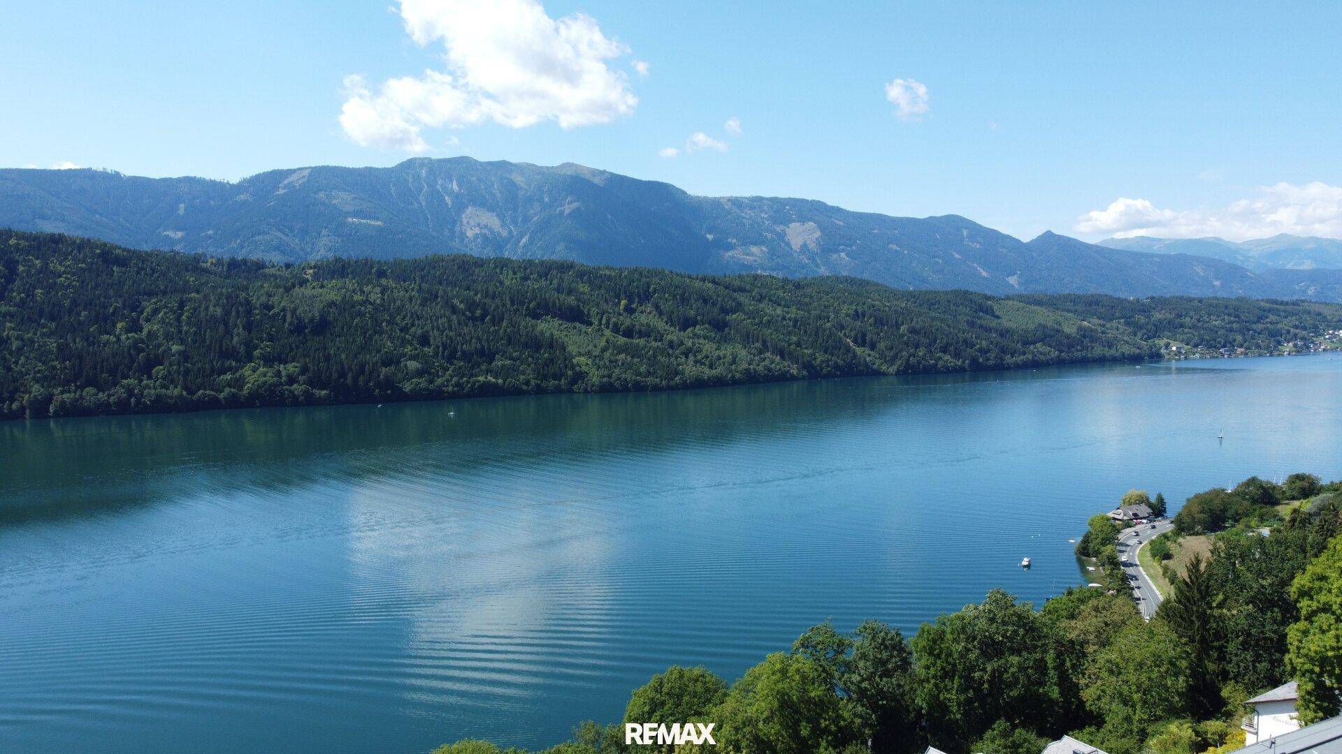 Weitläufiger Blick auf den See und die bewaldeten Berge unter einem klaren, blauen Himmel.