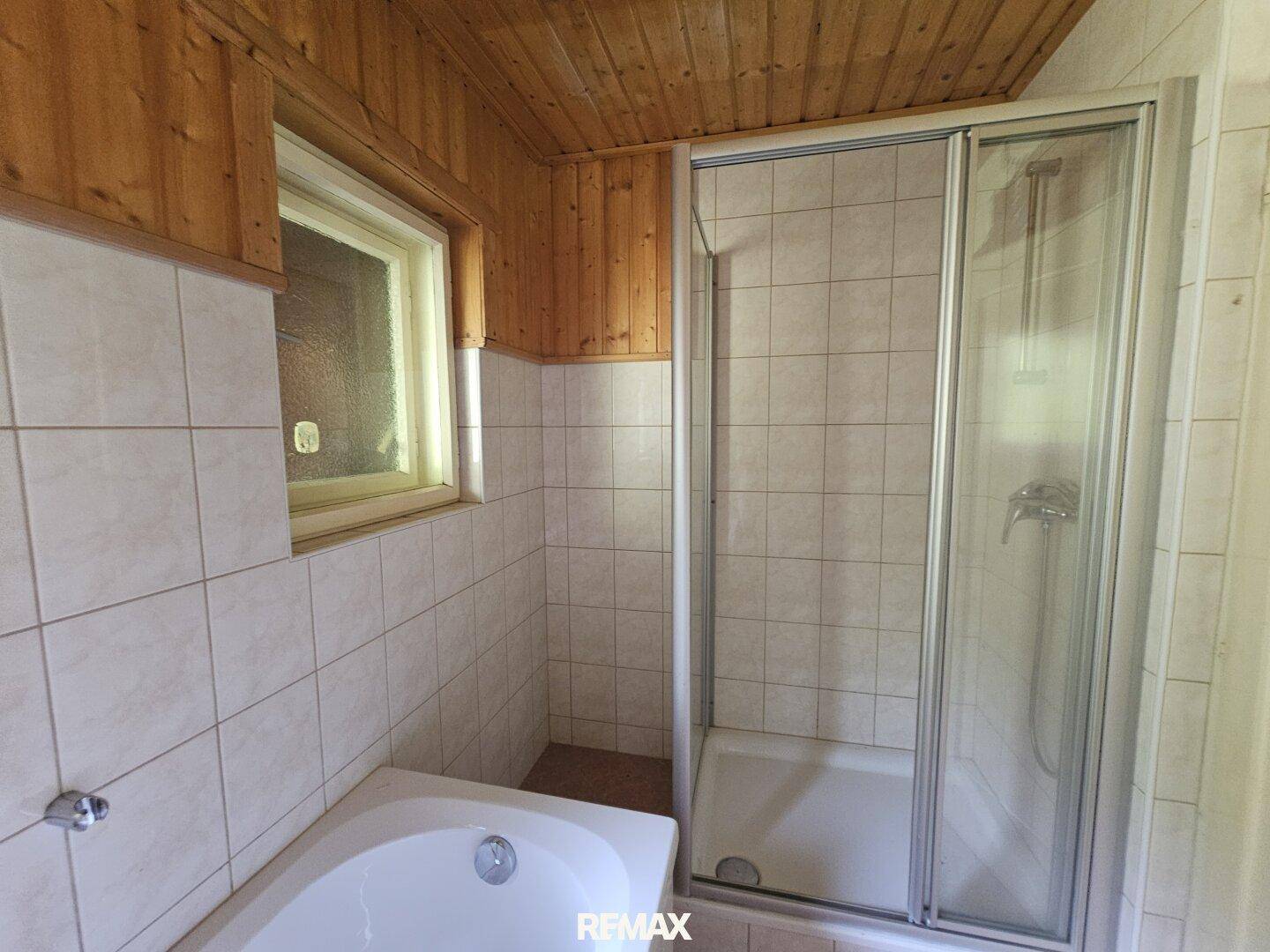 Badezimmer mit Duschkabine, hellen Fliesen und einer Holzdecke, gut beleuchtet durch ein Fenster.