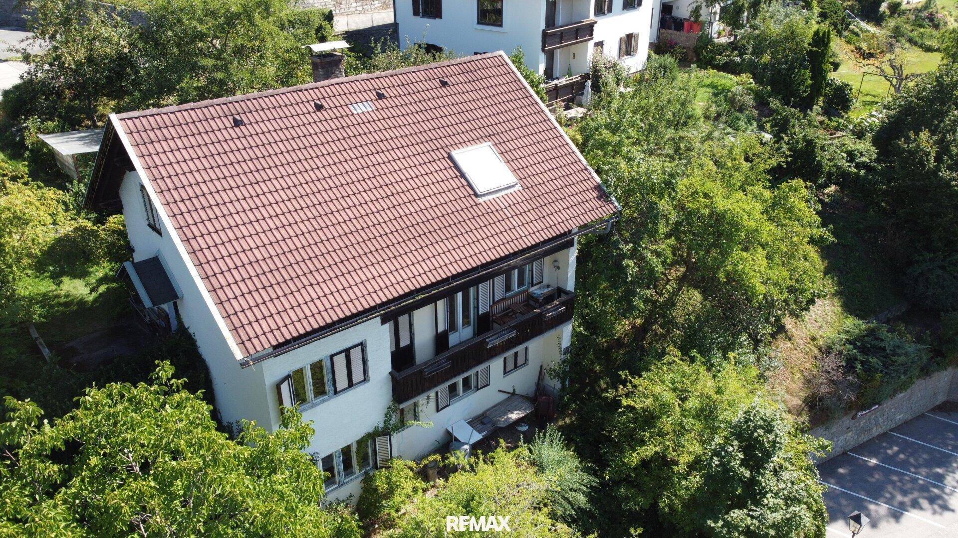 Das Haus mit rotem Ziegeldach und weißer Fassade, umgeben von Bäumen und einem kleinen Parkplatz.