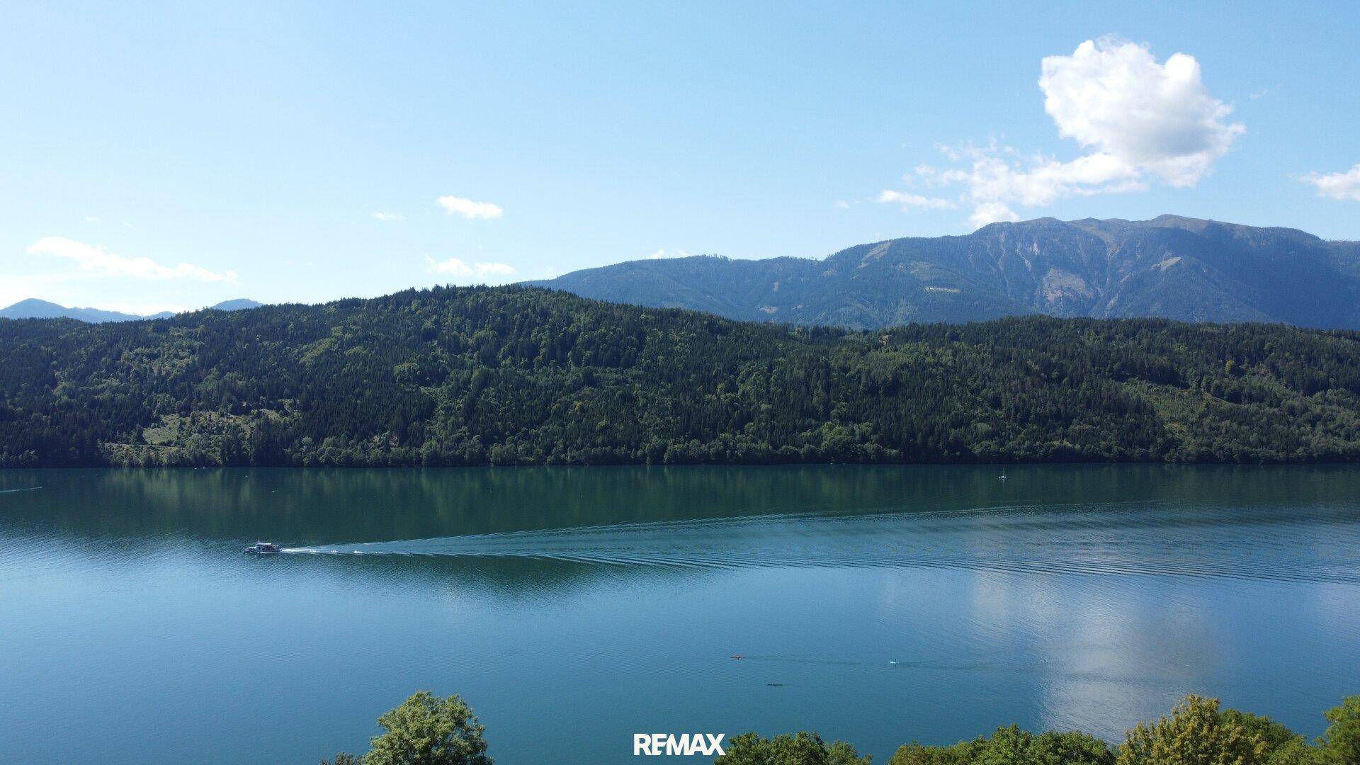 Panoramablick auf einen ruhigen See, umgeben von bewaldeten Bergen unter blauem Himmel.