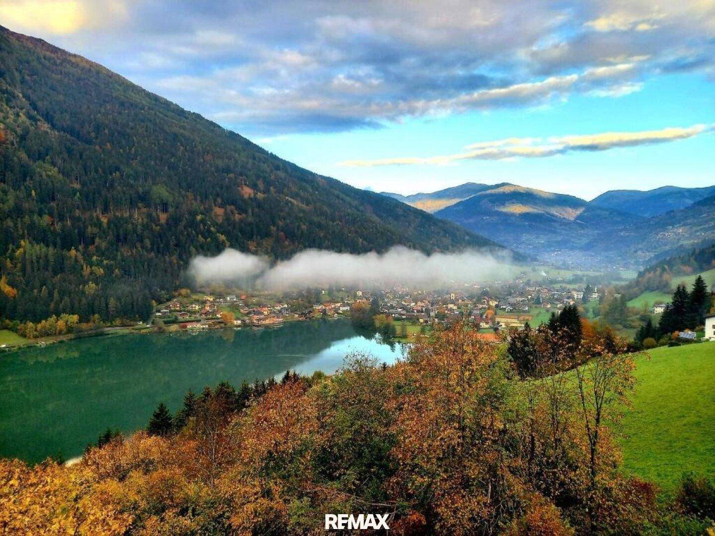 Herbstliche Berglandschaft mit einem See und Nebelschwaden, die über dem Dorf schweben.