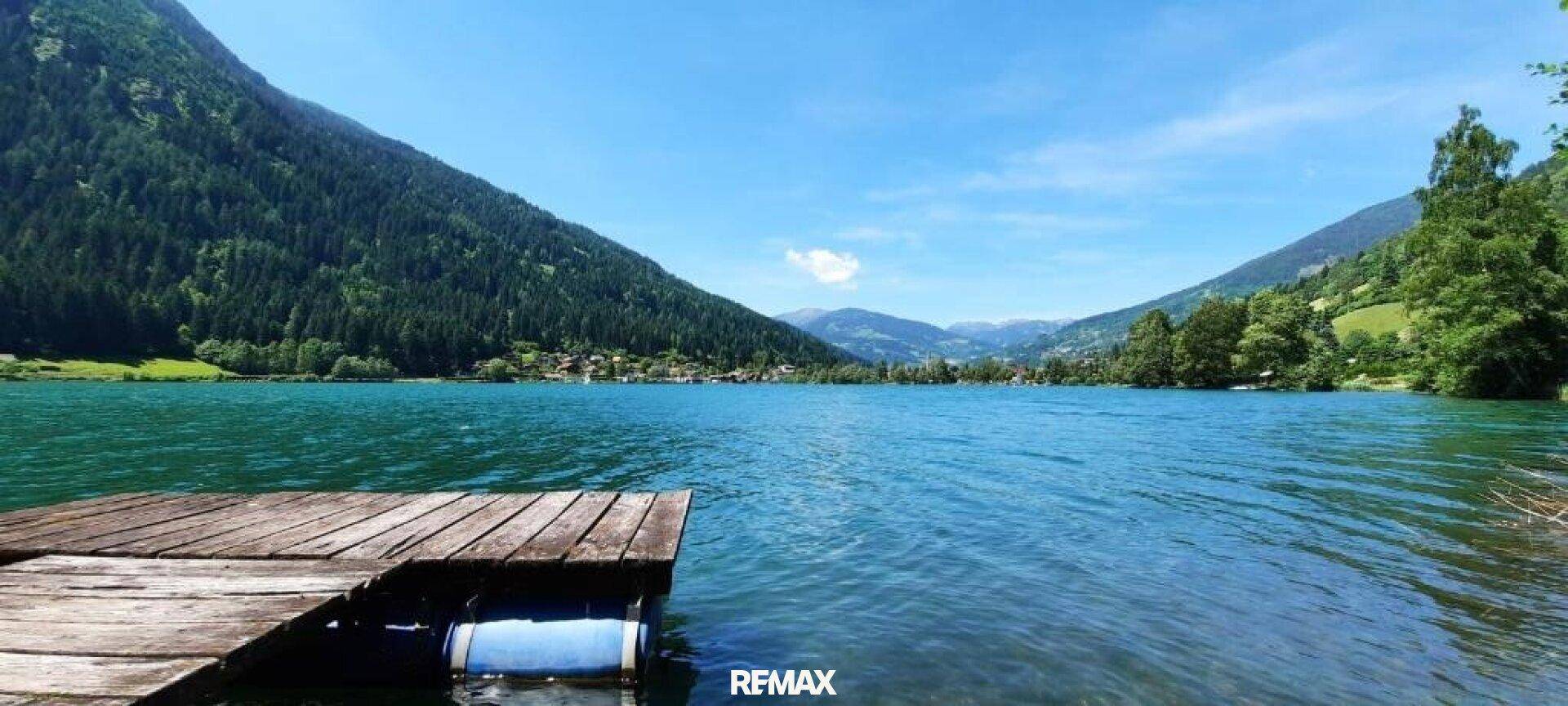 Idyllischer See mit Holzsteg, umgeben von grünen Bergen unter blauem Himmel.