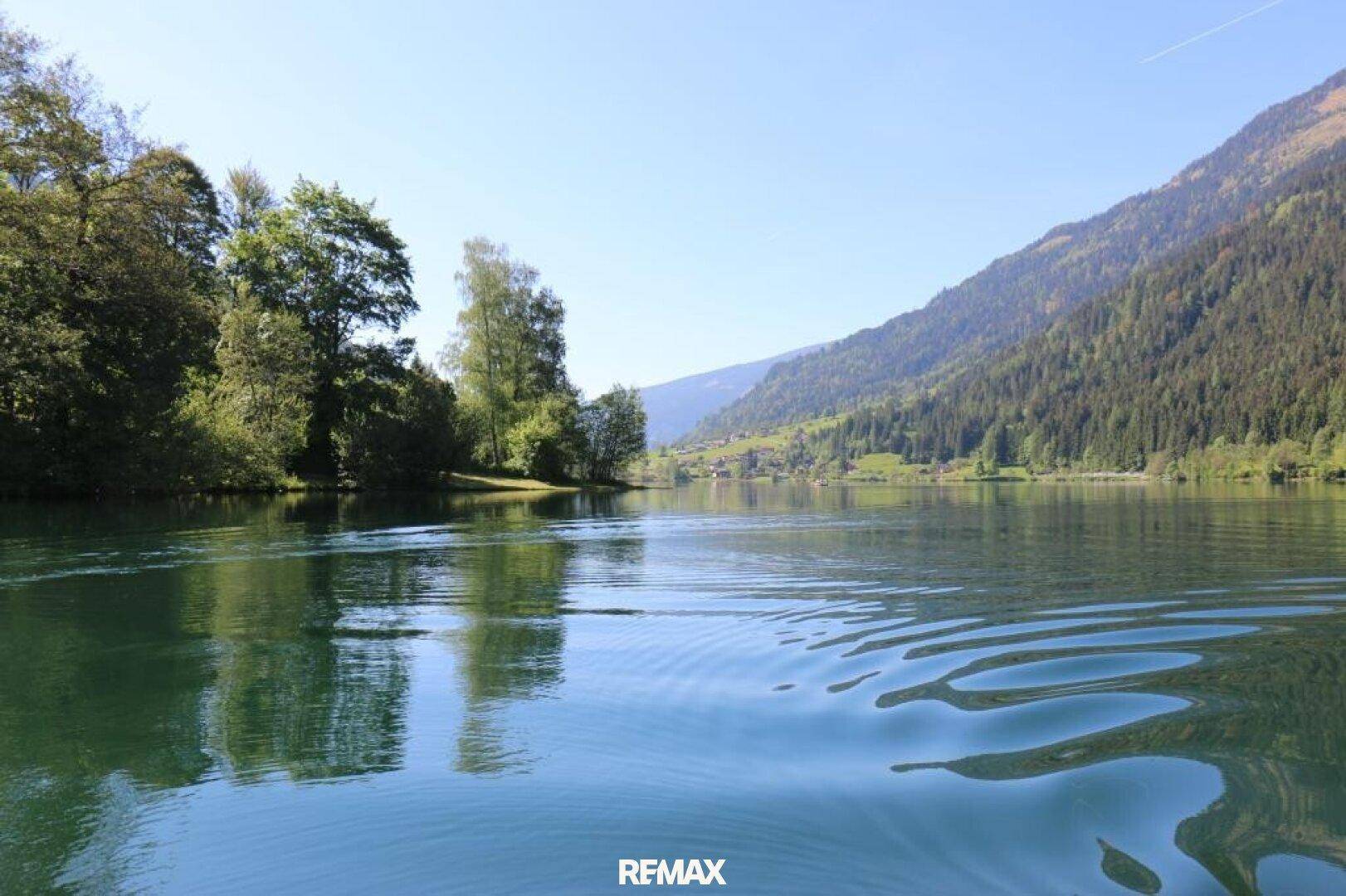 Ruhiger See mit spiegelnder Wasseroberfläche, umgeben von bewaldeten Bergen und Bäumen.