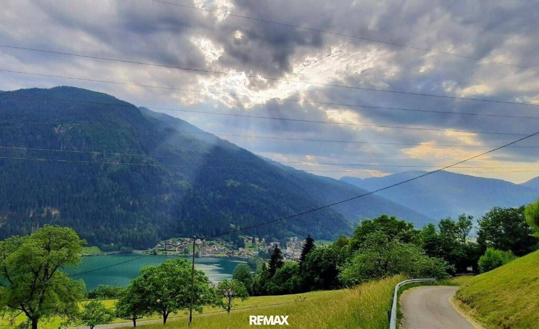 Malerische Landschaft mit einem See, umgeben von bewaldeten Bergen unter einem dramatischen Himmel.