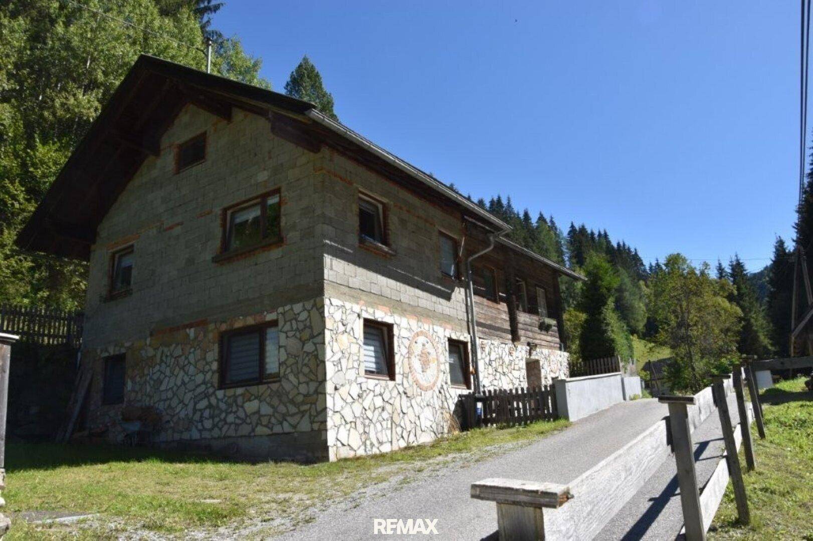 Ländliches Haus mit Steinfassade und Holzelementen in bergiger Umgebung unter blauem Himmel.