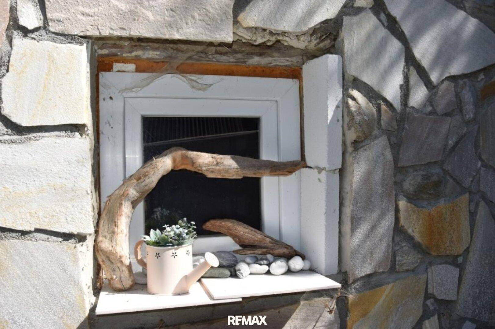 Kleines Fenster in einer Steinfassade, dekoriert mit Treibholz und Kieselsteinen.
