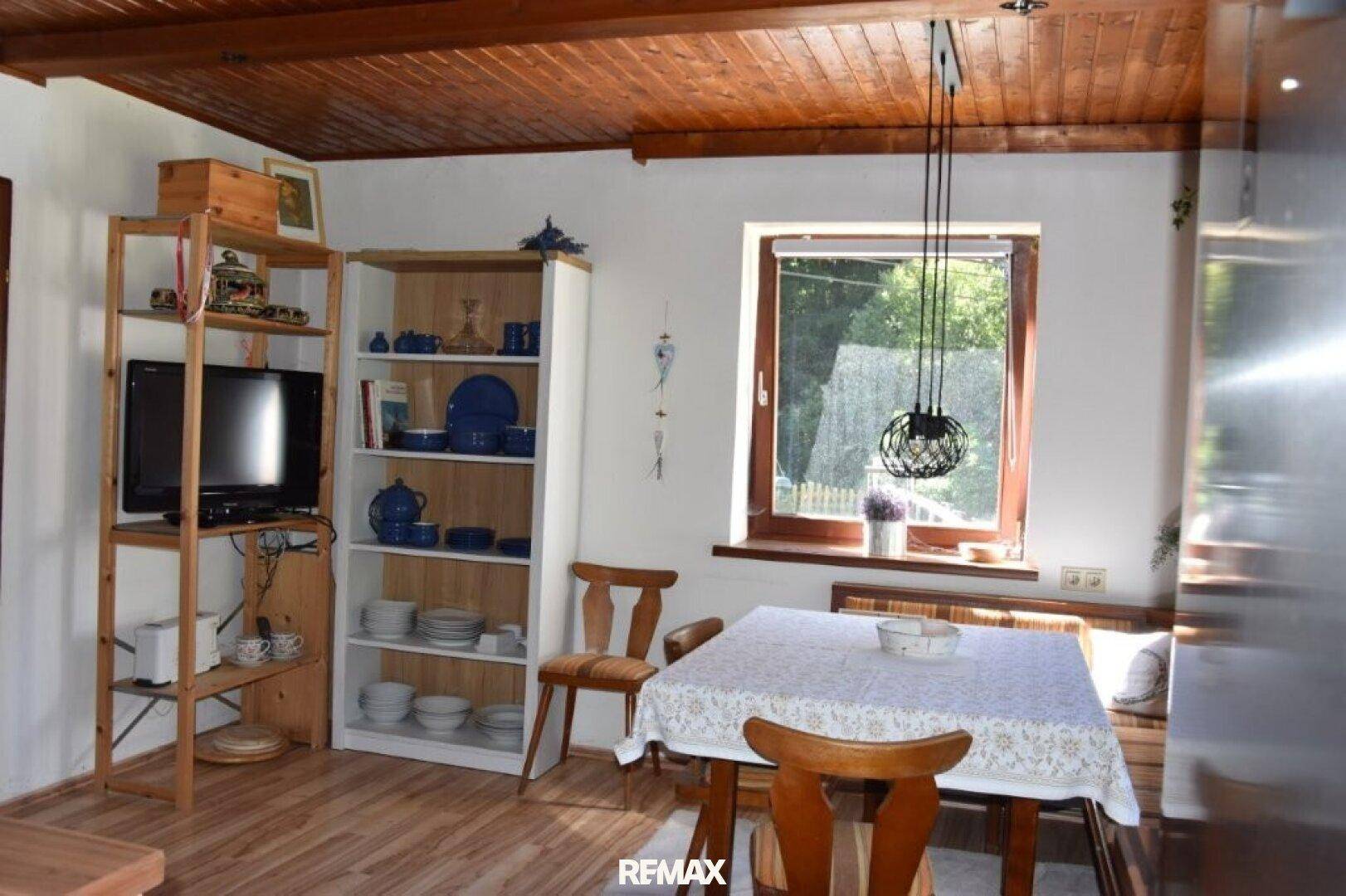 Essbereich mit Holztisch, Stühlen und Regal, beleuchtet durch ein Fenster mit Blick ins Grüne.