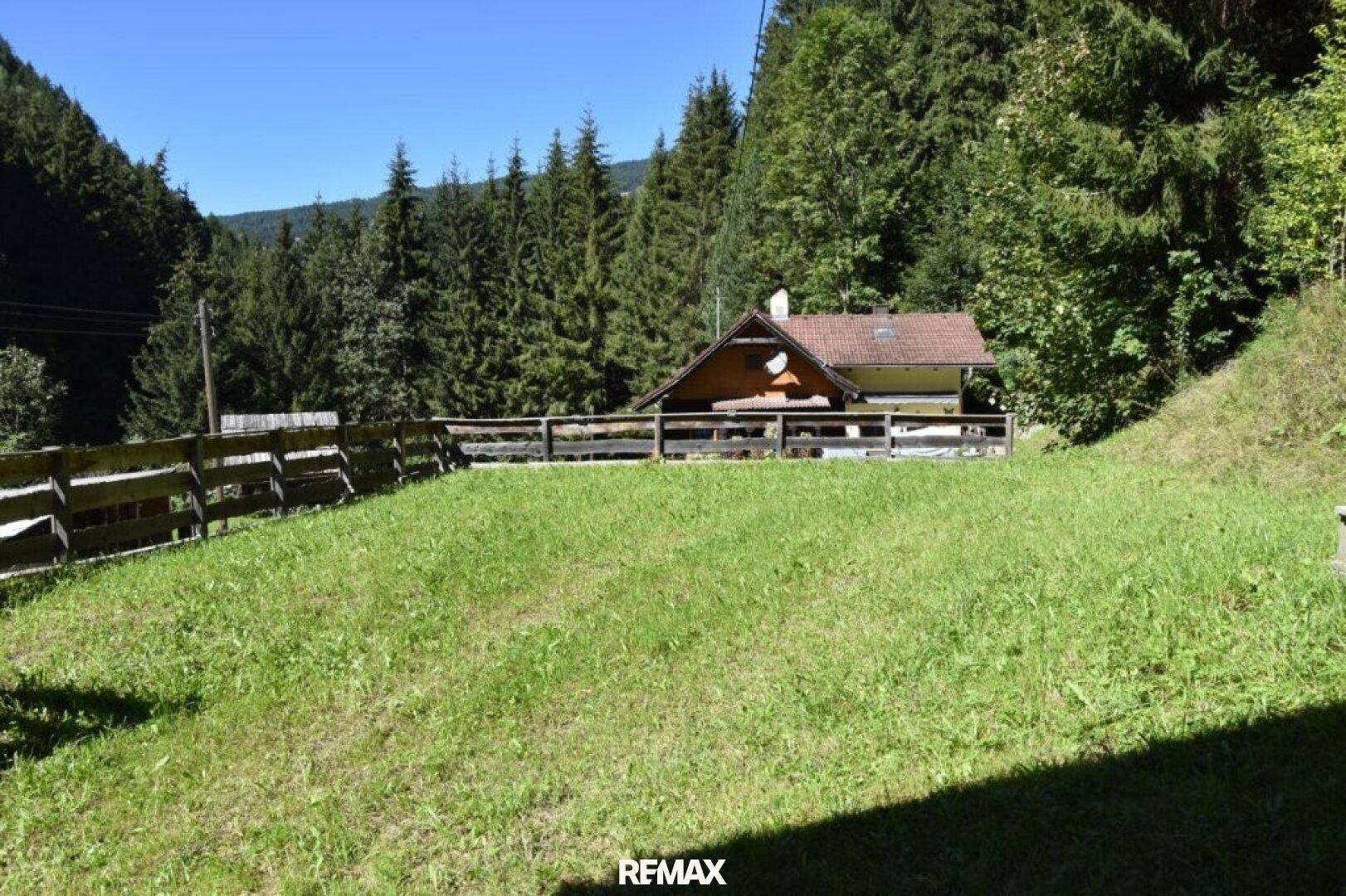 Grüne Rasenfläche mit Holzzaun und einem weiteren Gebäude im Hintergrund, umgeben von Wald.