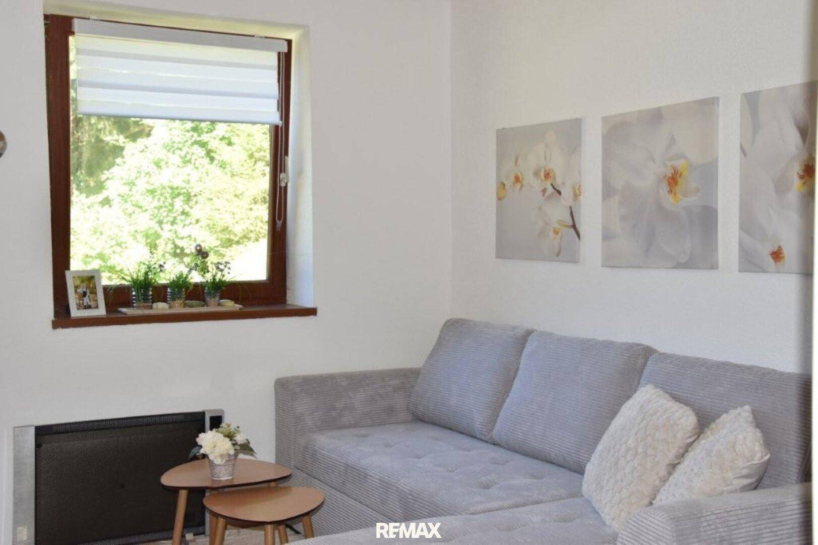 Helles Wohnzimmer mit grauem Ecksofa, Holzboden und Fenster mit Blick ins Grüne.