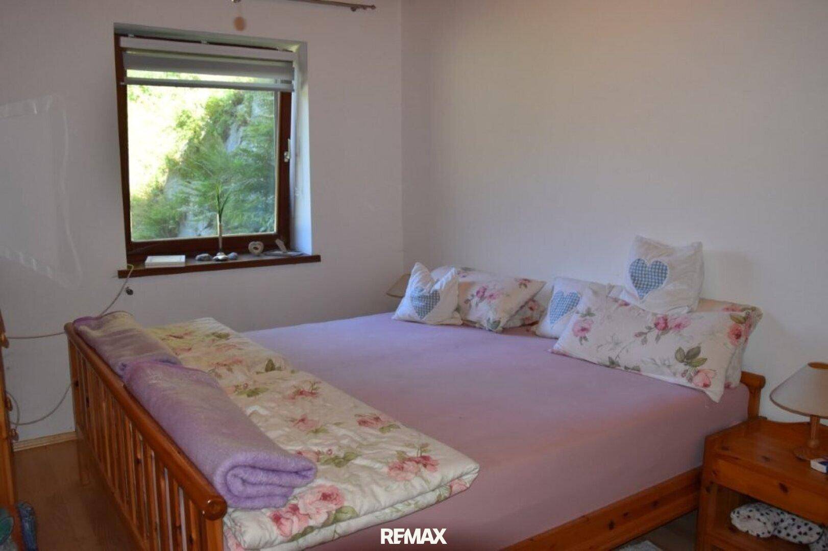 Helles Schlafzimmer mit Doppelbett, floralen Kissen und Fenster mit Blick ins Grüne.