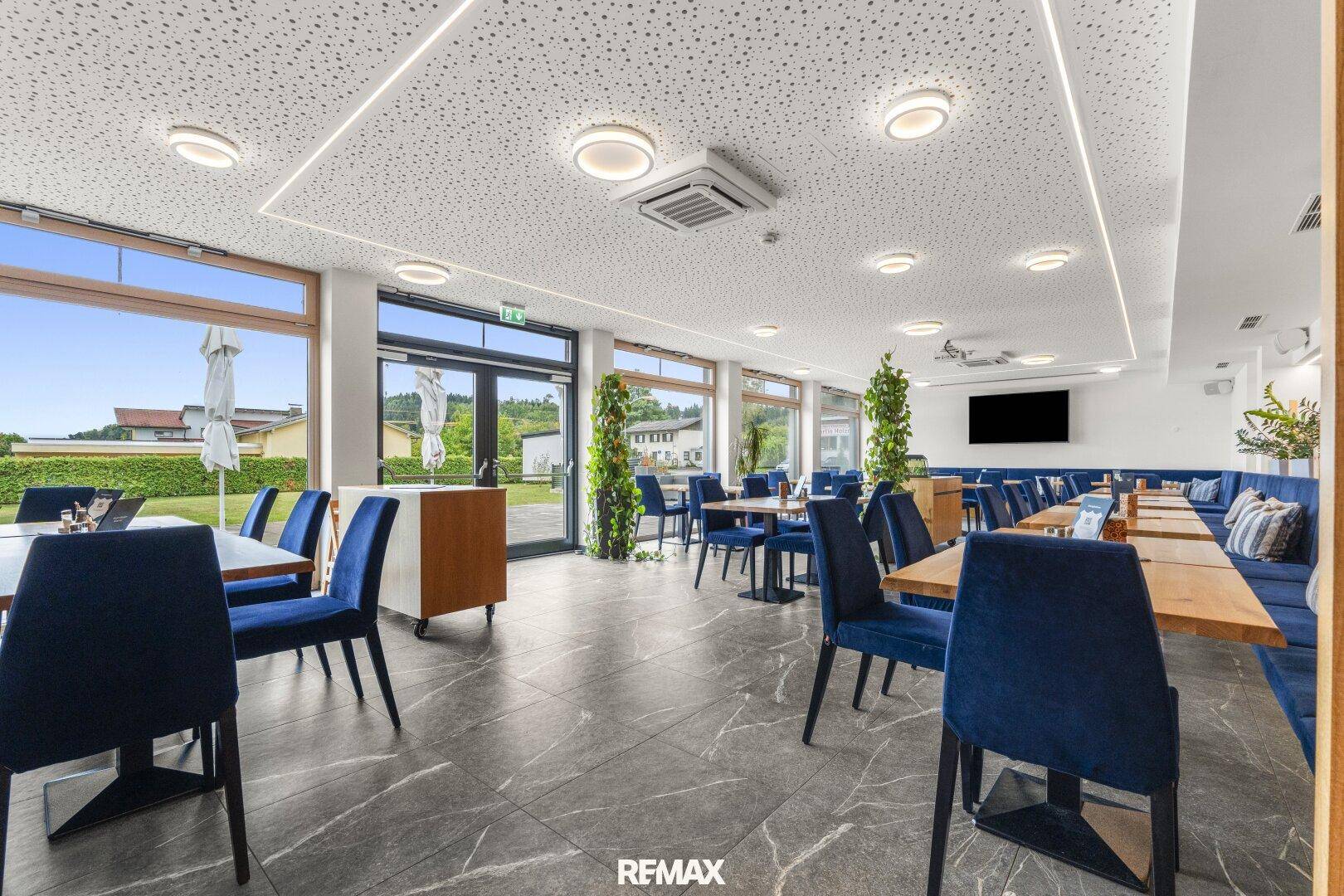 Heller Speisesaal mit blauen Stühlen, Holztischen und großen Fenstern mit weitem Blick ins Grüne.