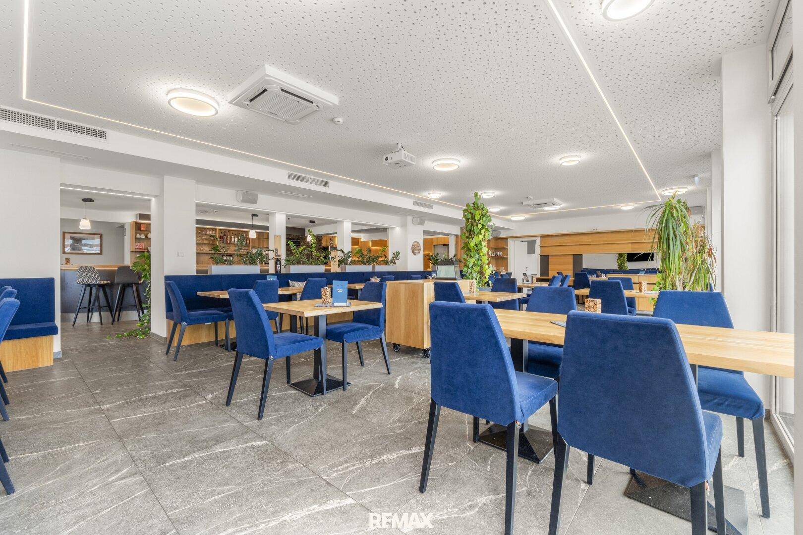 Geräumiger Speisesaal mit blauen Sitzgelegenheiten, Holztischen und offenem Übergang zum Barbereich im Hintergrund.