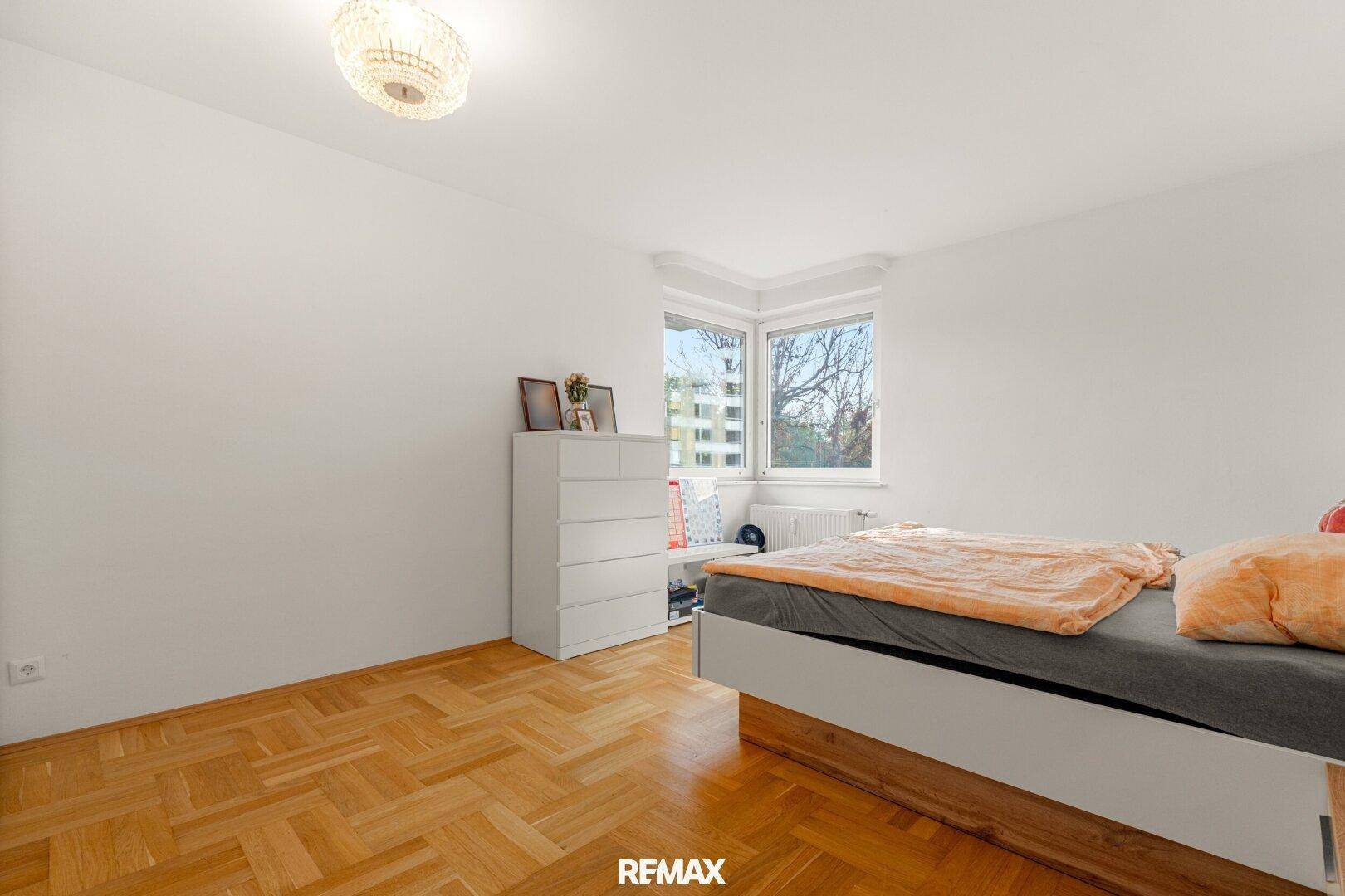 Helles Schlafzimmer mit Parkettboden, weißer Kommode und großem Fenster.