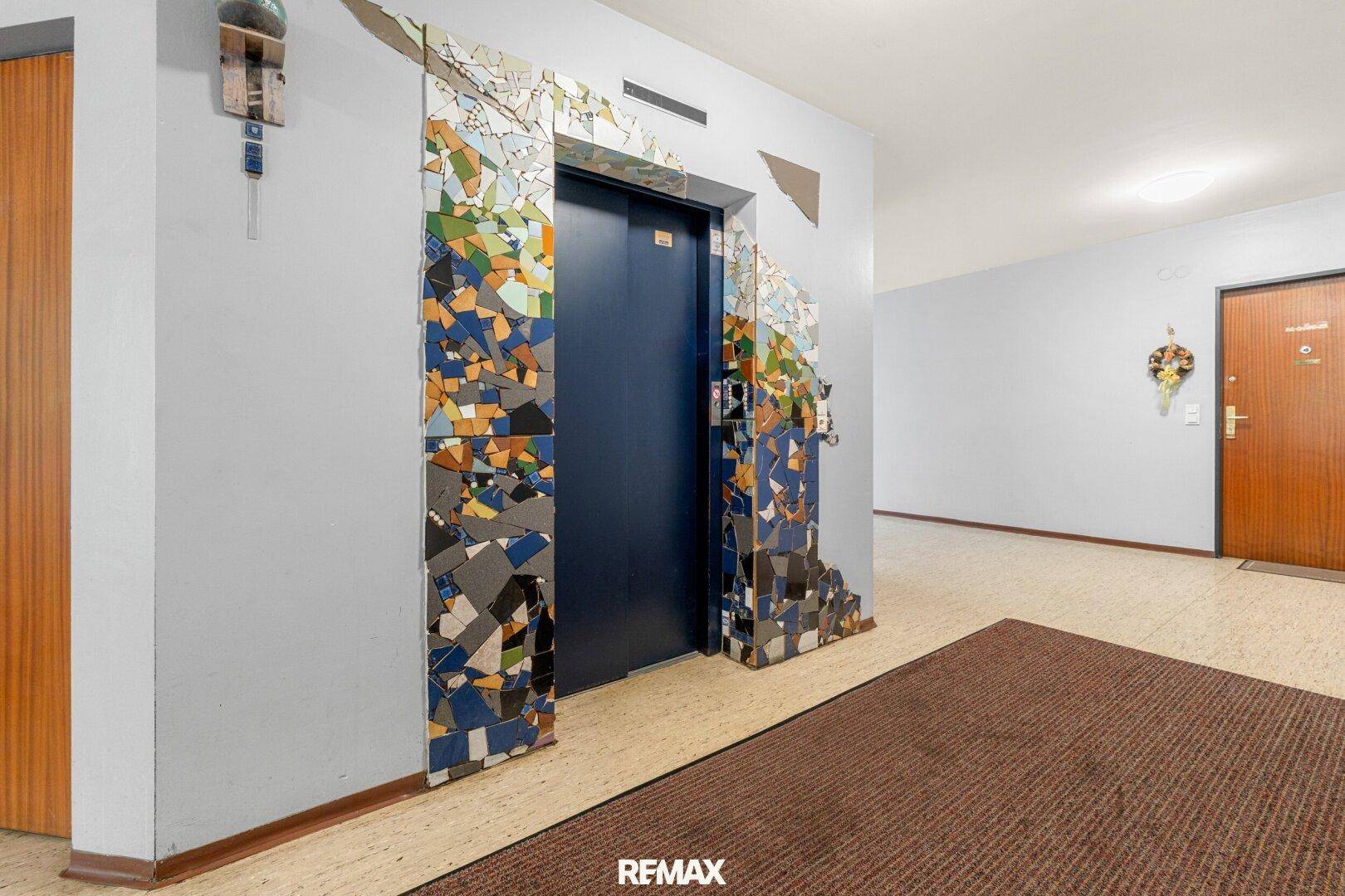 Eingangsbereich mit Aufzug und farbenfroher Mosaikverzierung an der Wand.