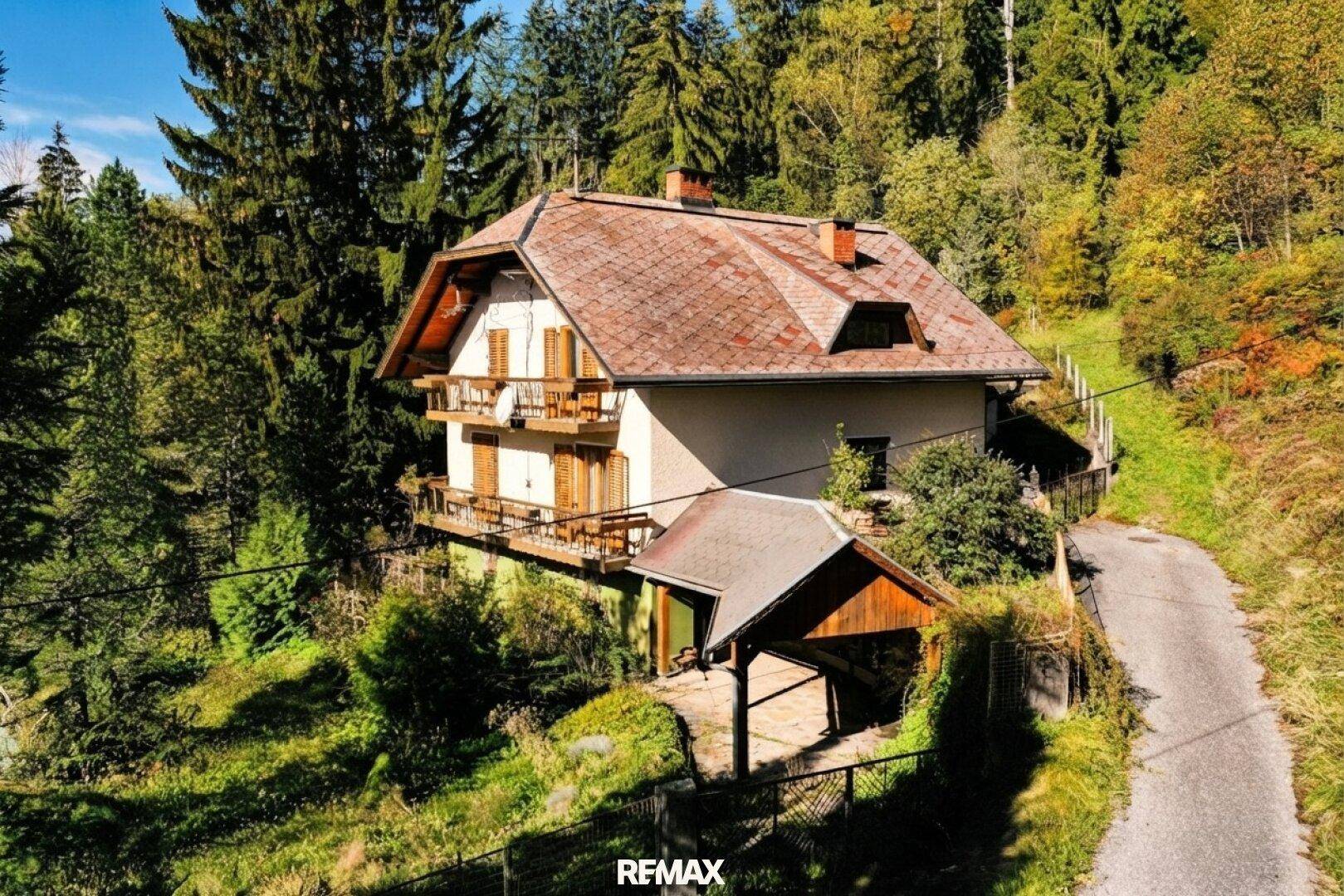 Charmantes Haus mit Satteldach und Holzbalkonen, eingebettet in eine grüne Hügellandschaft mit Wald.