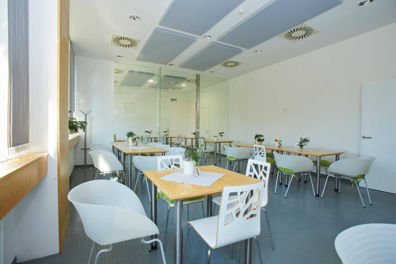 Helle Cafeteria mit vielen Tischen und Stühlen, großen Fenstern und schallabsorbierenden Deckenplatten.