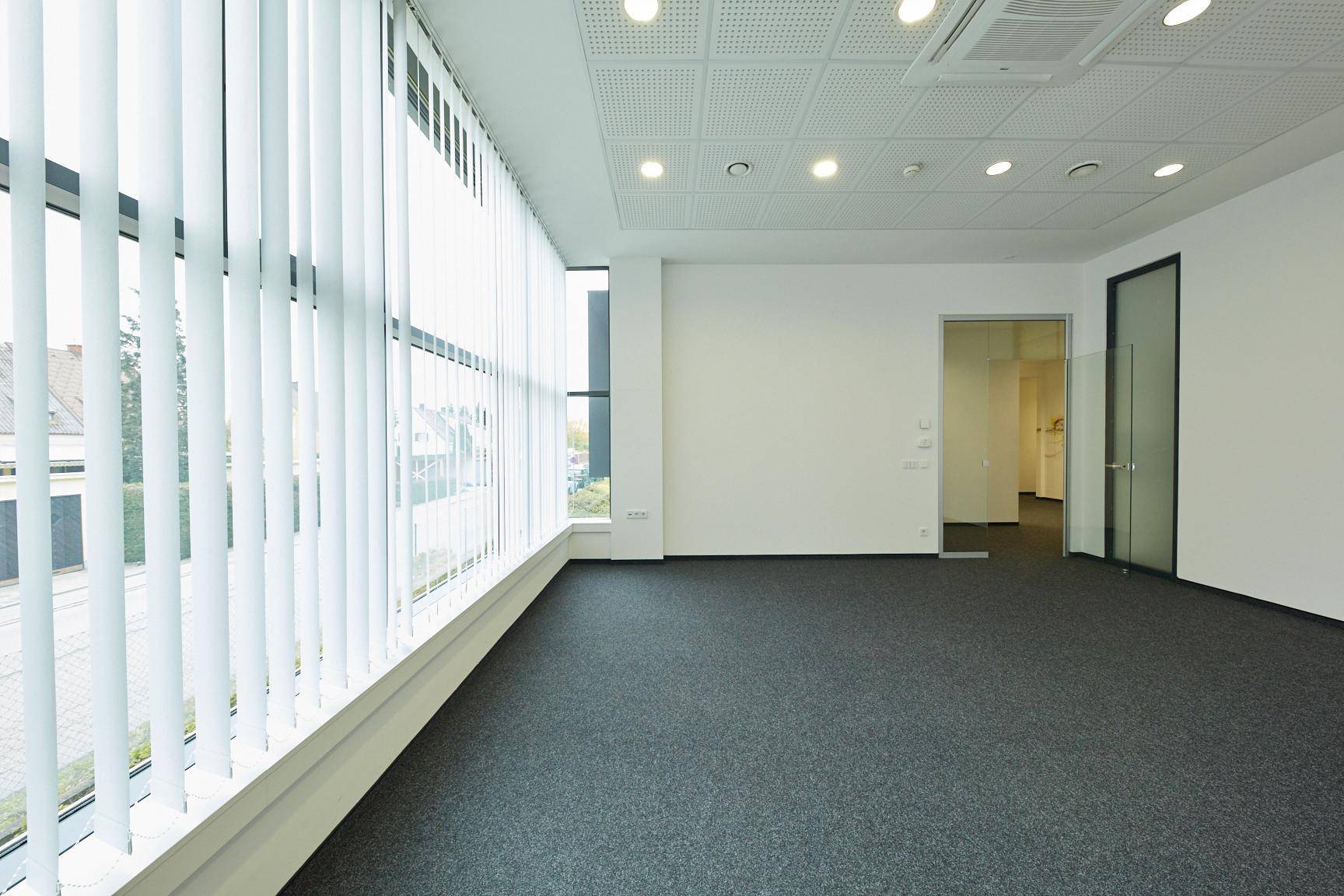 Geräumiger, leerer Büroraum mit großen Fenstern, vertikalen Jalousien und grauem Teppichboden.