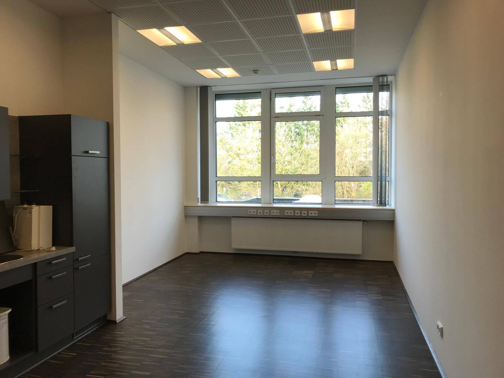 Heller Büroraum mit dunklem Boden, weißen Wänden und einer sichtbaren Küchenzeile am Rand.