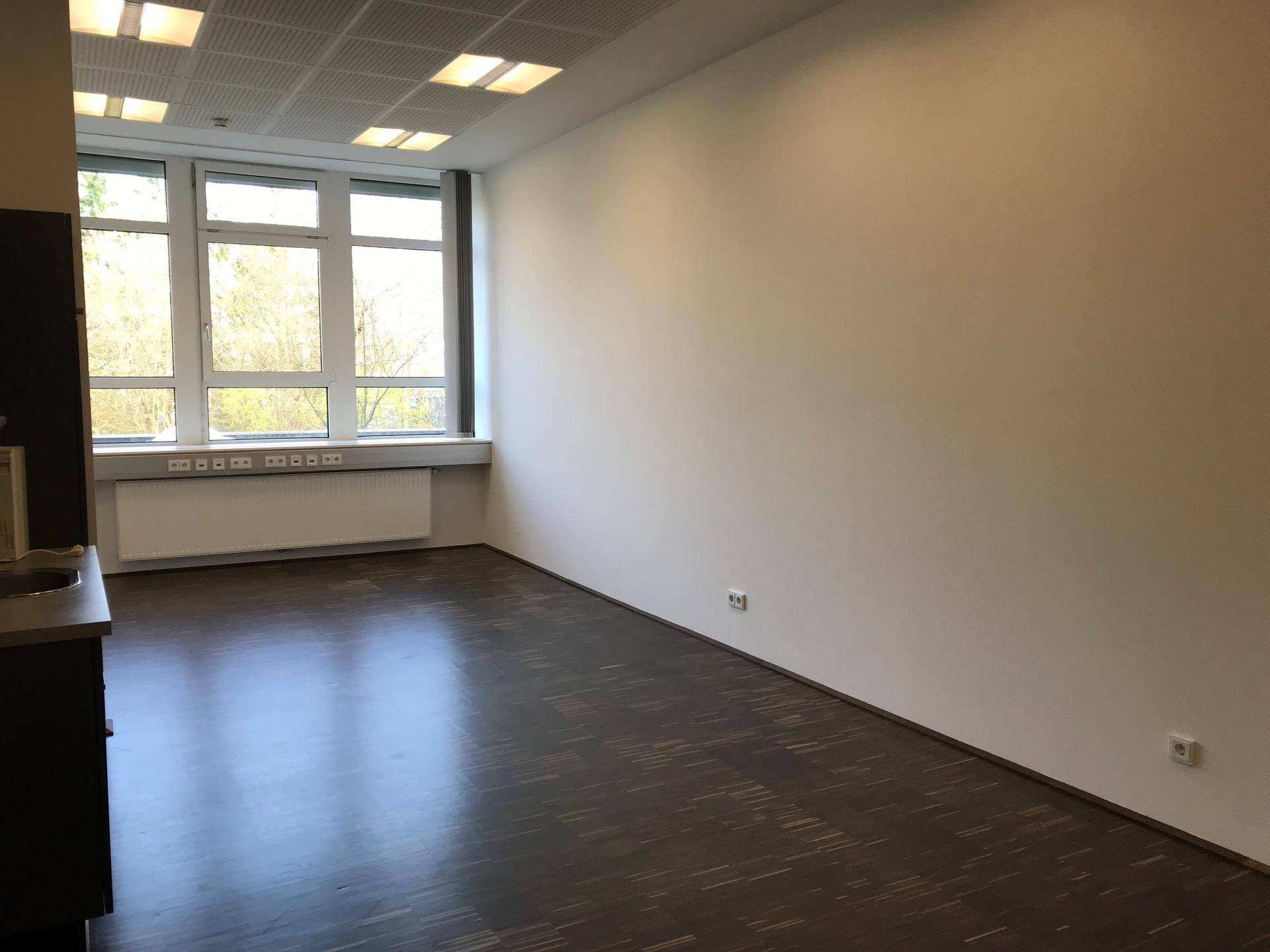 Leerer Büroraum mit dunklem Holzboden, weißen Wänden und großen Fenstern, die viel Tageslicht hereinlassen.