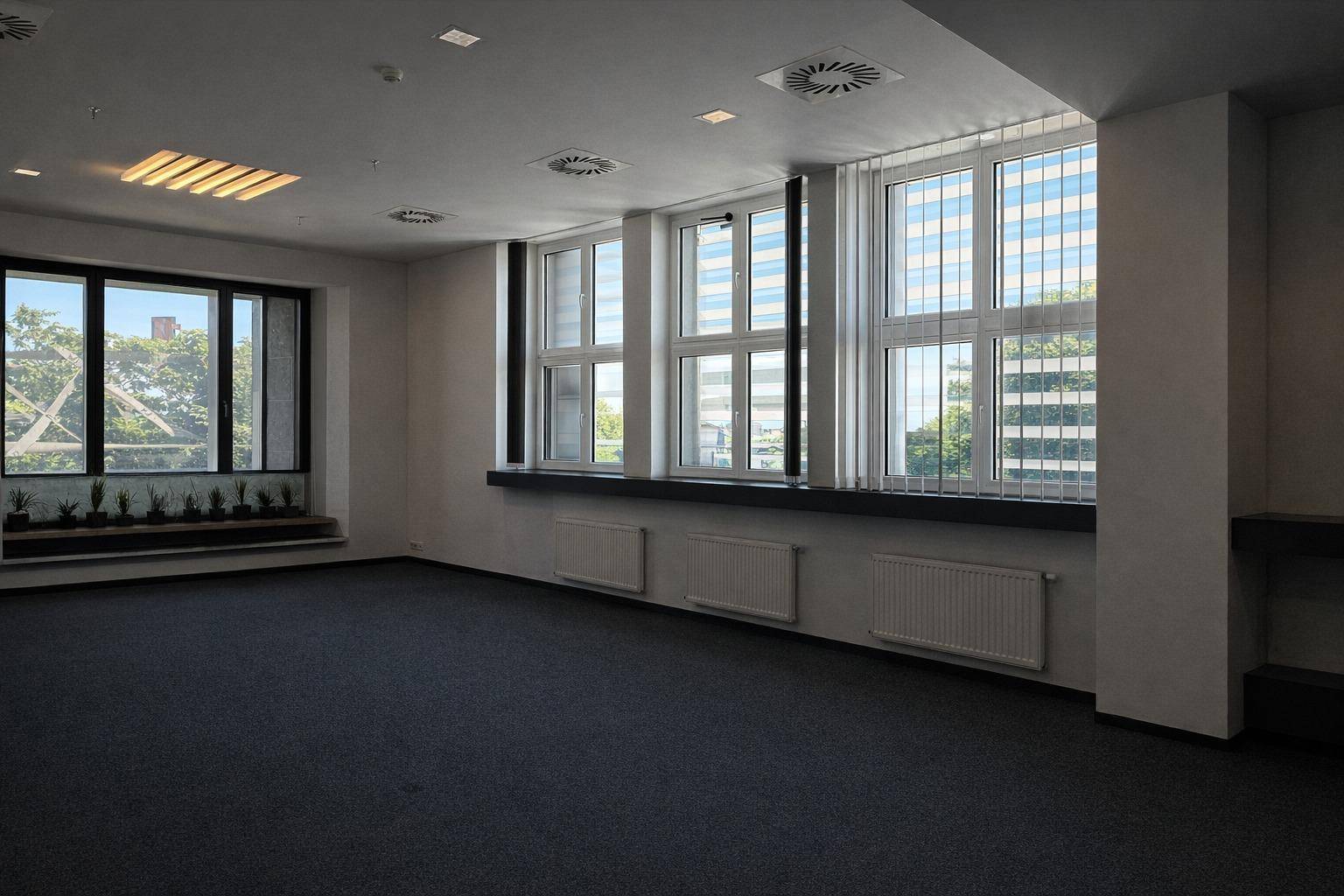 Leerer Büroraum mit dunklem Teppichboden, großen Fenstern und Lüftungsanlagen an der Decke.
