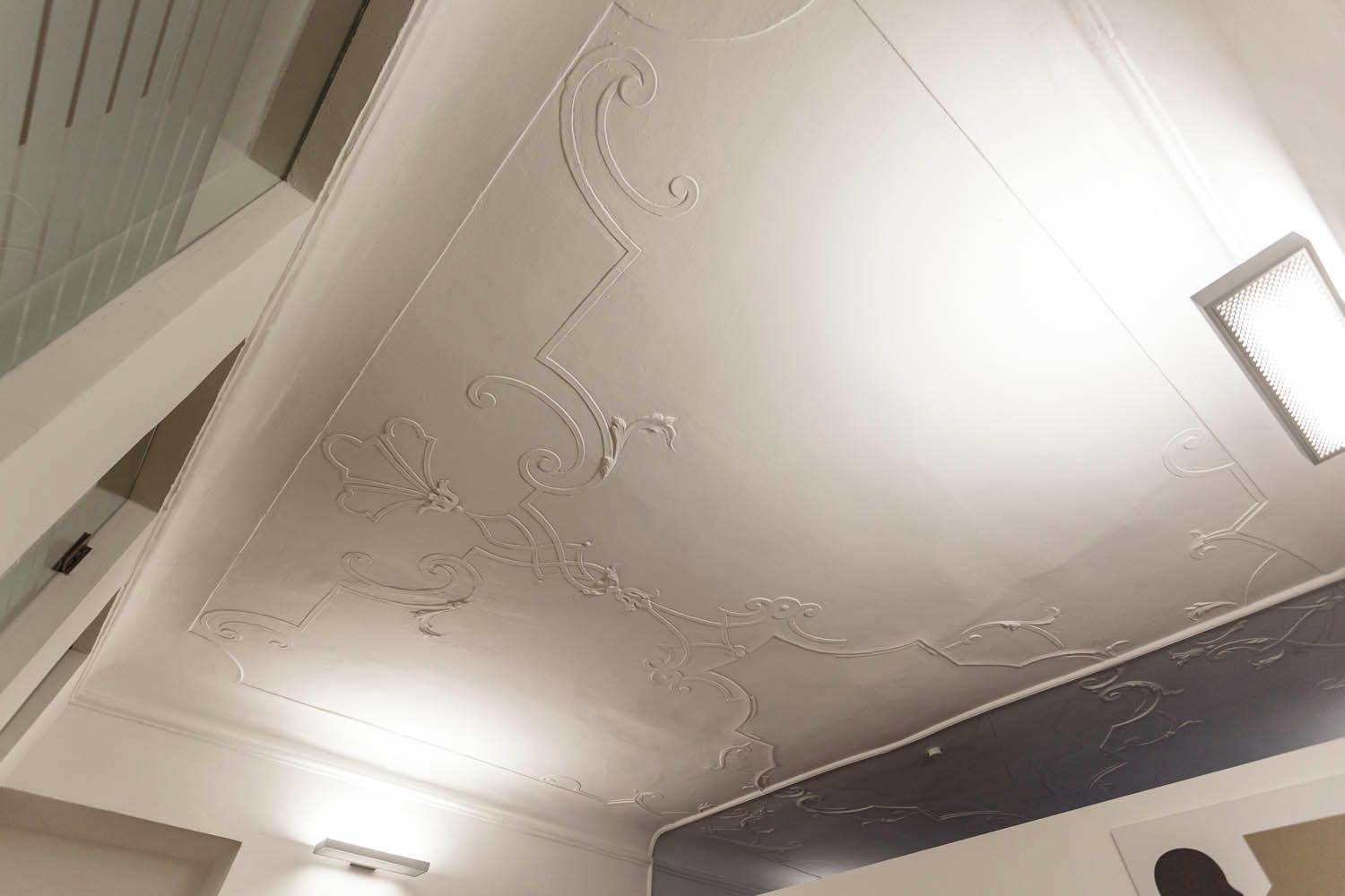 Weiße Decke mit kunstvollen Stuckverzierungen und einer modernen Beleuchtung, die das Muster hervorhebt.