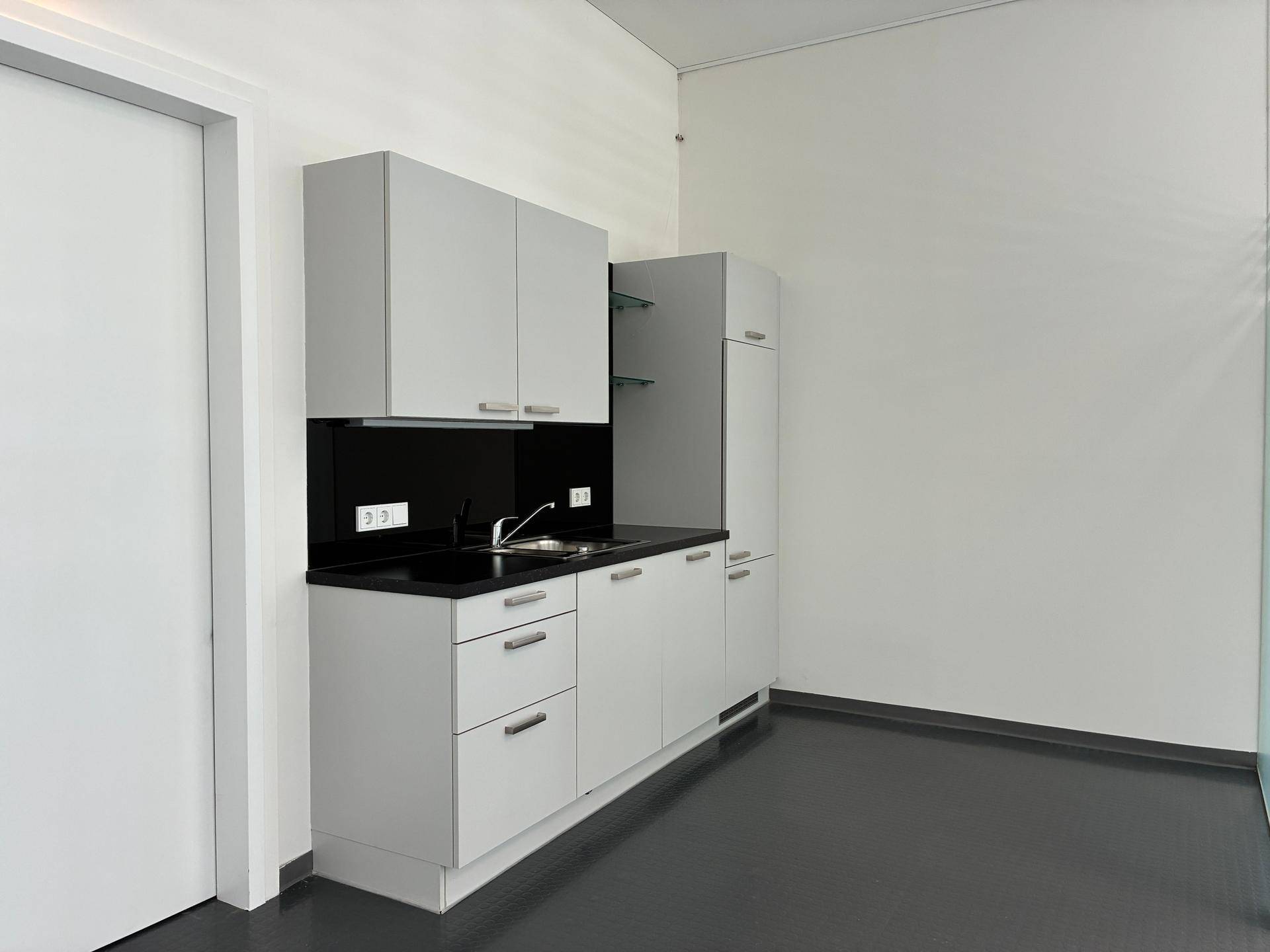 Kleine, funktionale Küchenzeile mit grauen Schränken, schwarzer Arbeitsplatte und Spülbecken in einem hellen Raum.