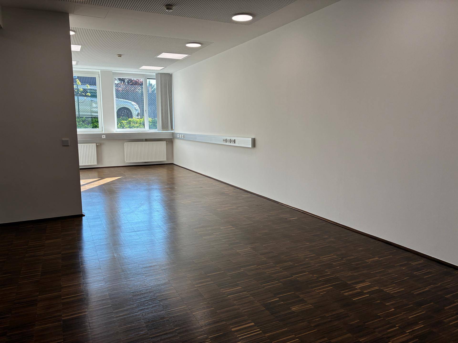 Geräumiges, leeres Büro mit Holzboden und großen Fenstern, das viel natürliches Licht bietet.