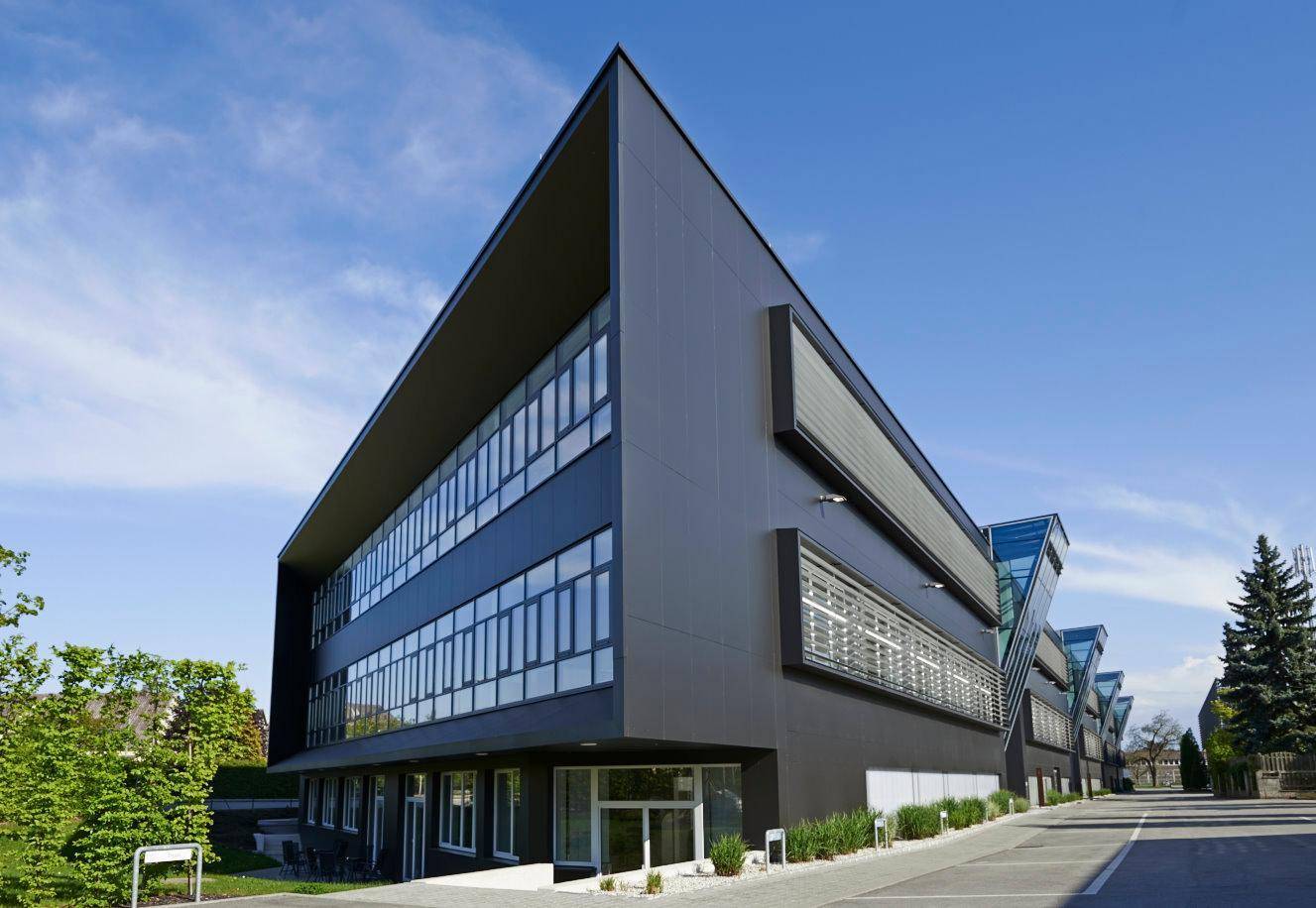 Modernes Bürogebäude mit dunkler Fassade, großen Fenstern und markanter Architektur unter blauem Himmel.