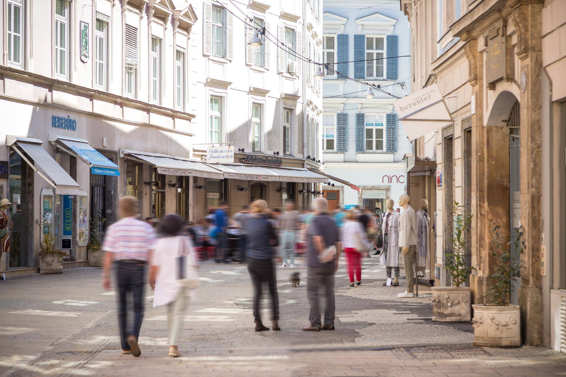 Belebte Einkaufsstraße mit vielen Passanten, Geschäften und historischen Gebäuden unter blauem Himmel.
