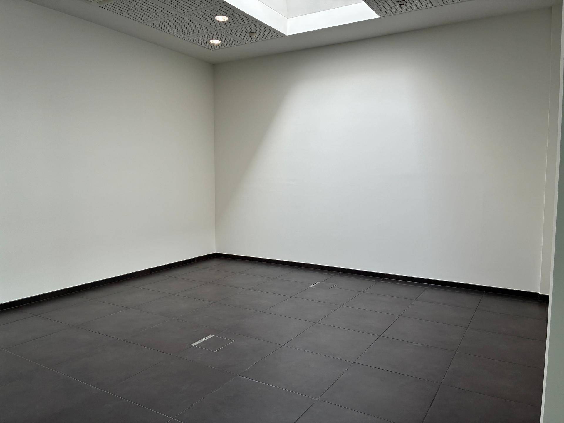 Leerer Raum mit dunklem Fliesenboden, weißen Wänden und einem Oberlicht für Tageslicht.