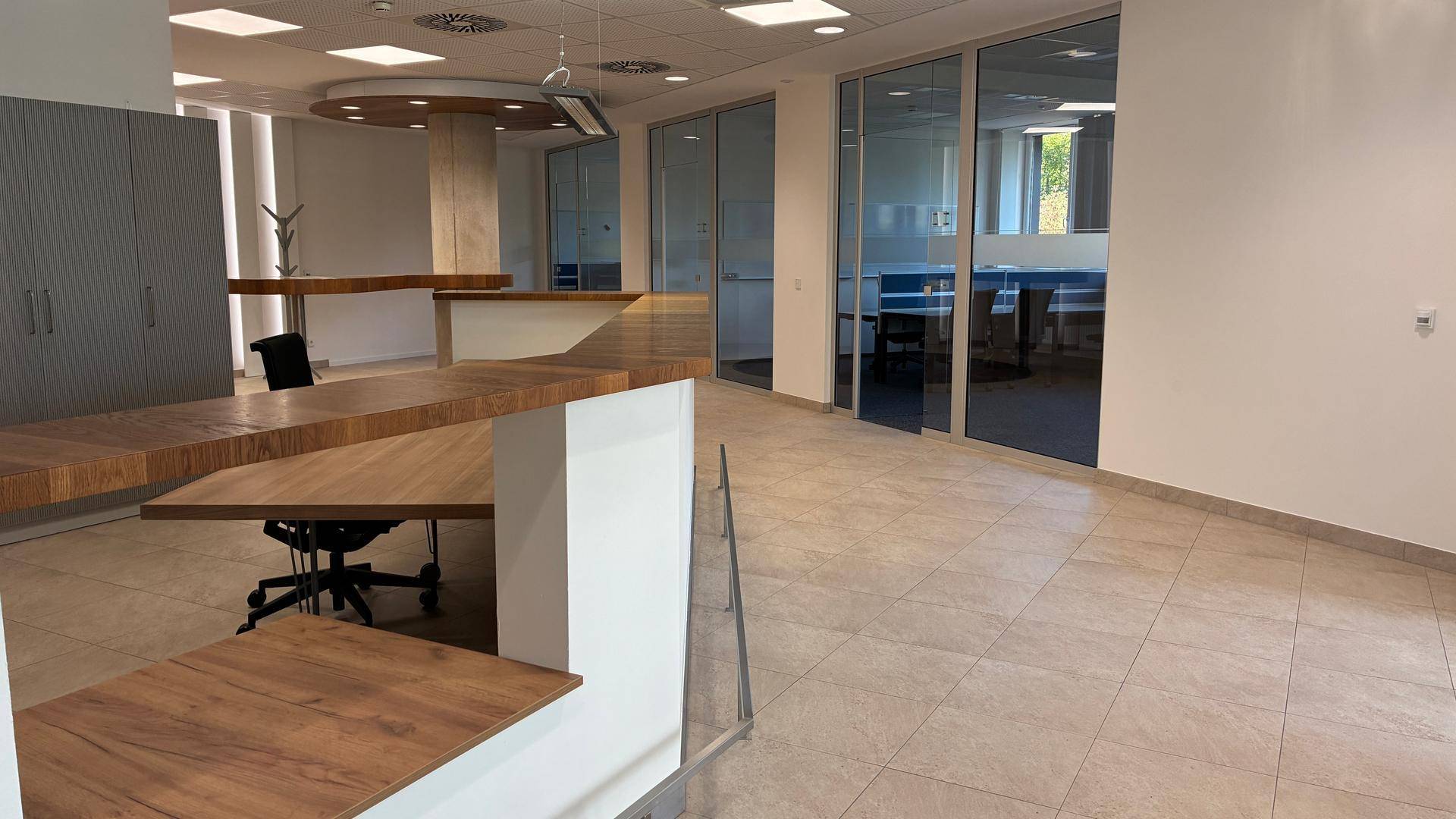 Moderner Bürobereich mit hellen Fliesenböden, Holztischen und Glaswänden, die separate Arbeitsbereiche abgrenzen.